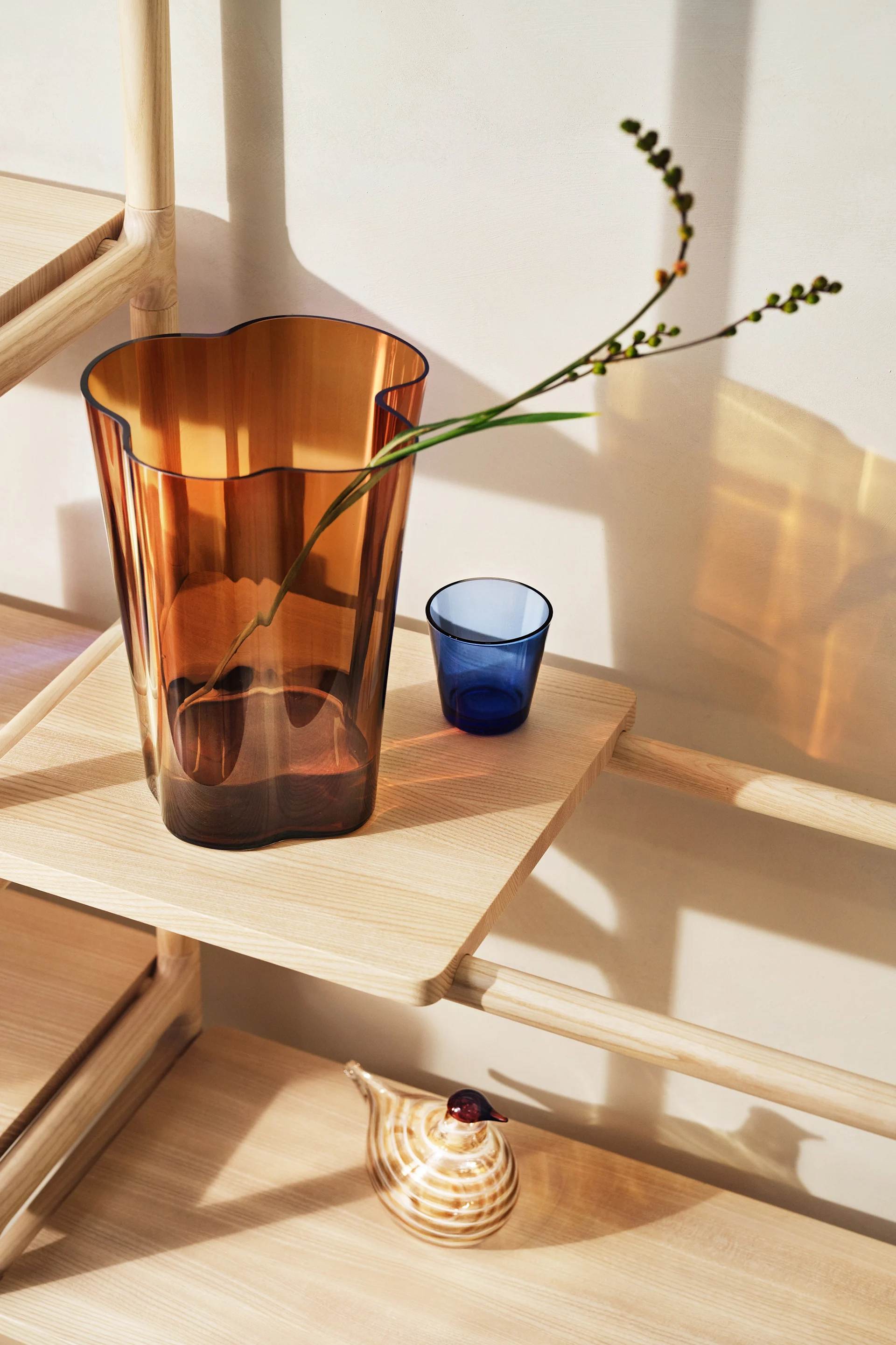 Jarrón ámbar con rama, vaso azul y pájaro de cristal a rayas en una estantería de madera clara.