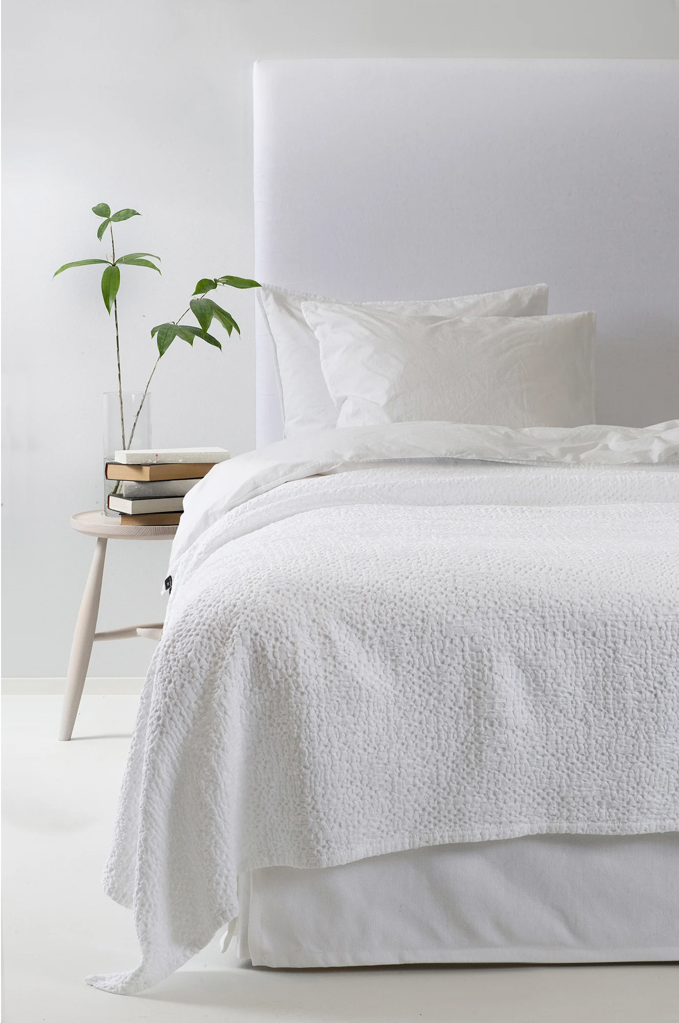 Cama blanca con edredón texturizado, almohadas, cabecero tapizado, mesita de madera con libros y planta verde.