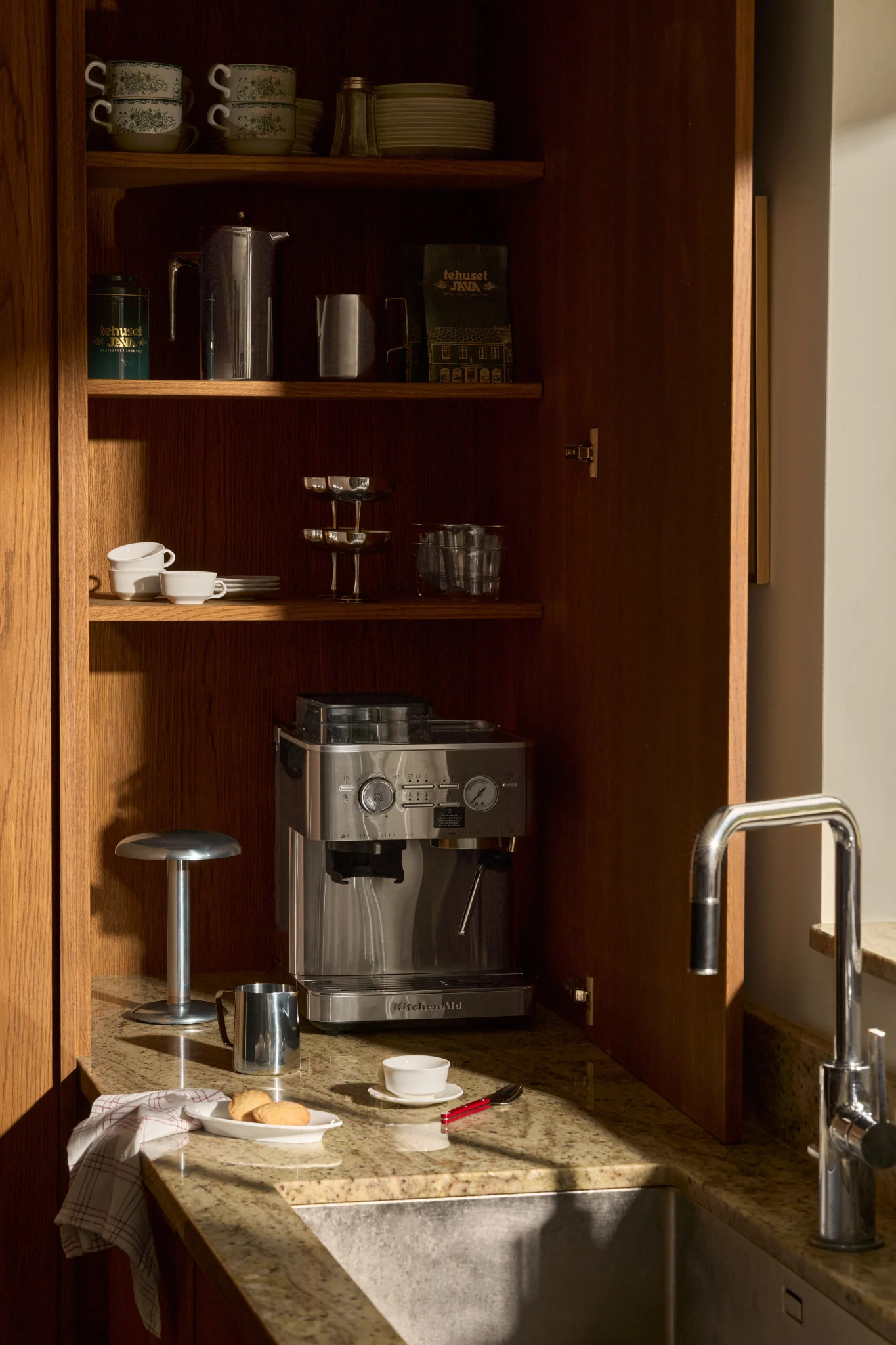 Un armario de cocina de madera abierto con cafetera espresso, accesorios de café, tazas apiladas y vasos en estantes, junto a un fregadero.
