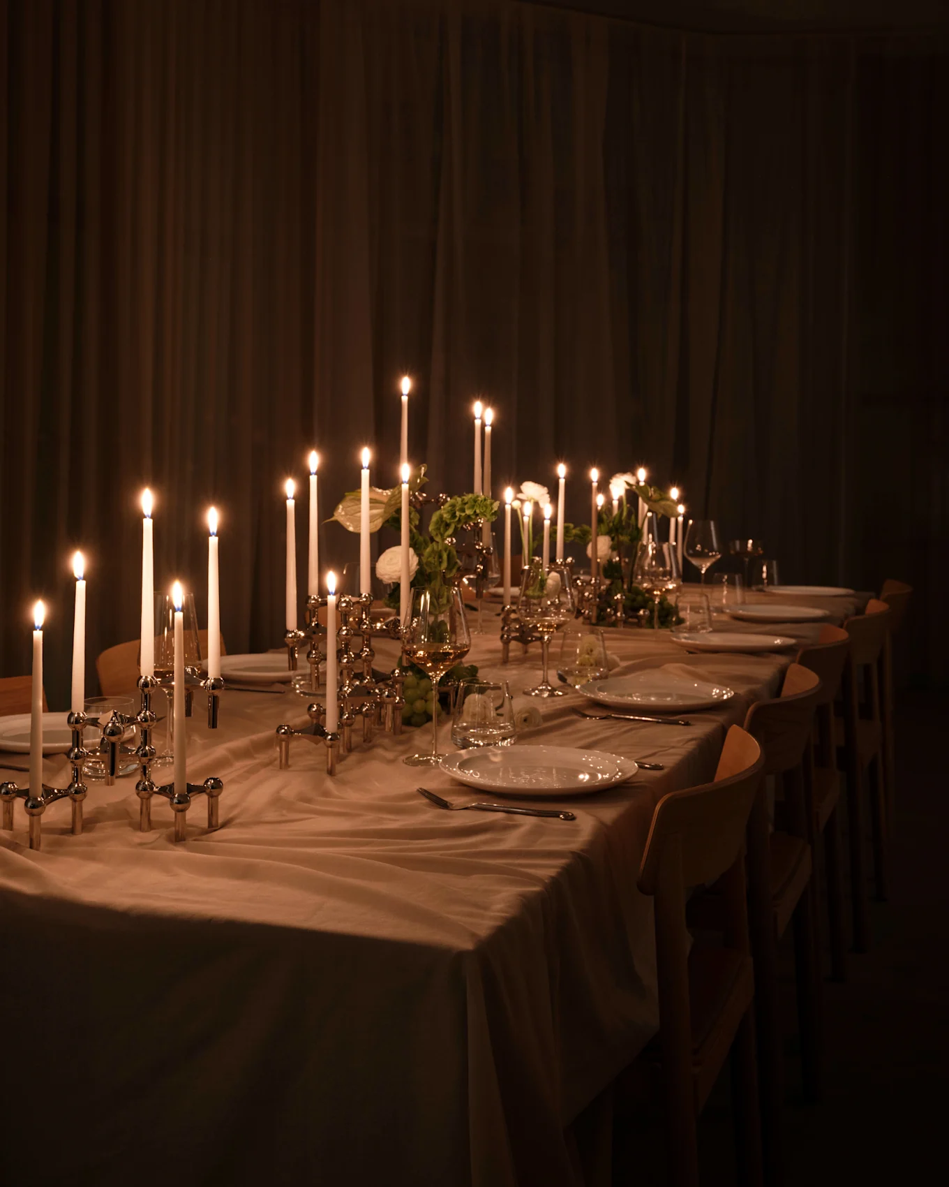 Larga mesa de comedor con velas encendidas en candelabros plateados, platos blancos y vasos, luz cálida en habitación tenue.