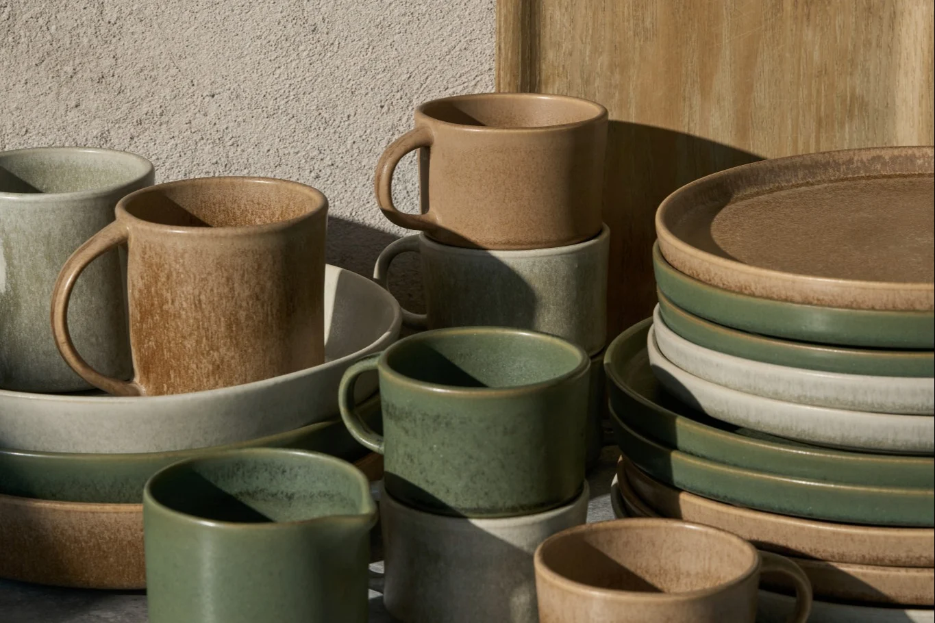 Tazas, cuencos y platos de cerámica apilados en beige, gris claro y verde salvia, con acabado rústico.