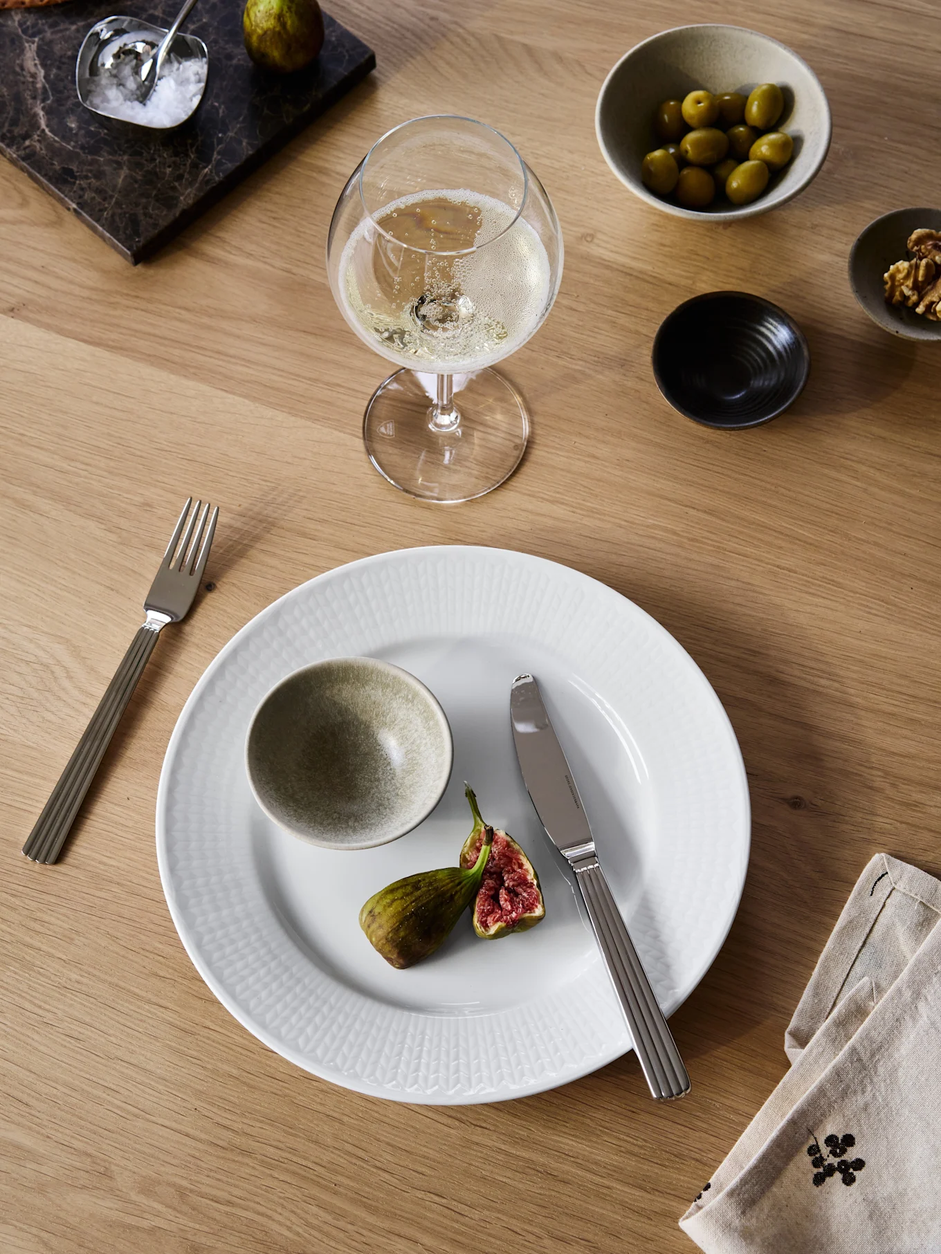 Mesa de comedor con plato blanco, higo partido, tenedor, cuchillo, vino espumoso, aceitunas, nueces, galletas saladas y sal sobre tabla de mármol.