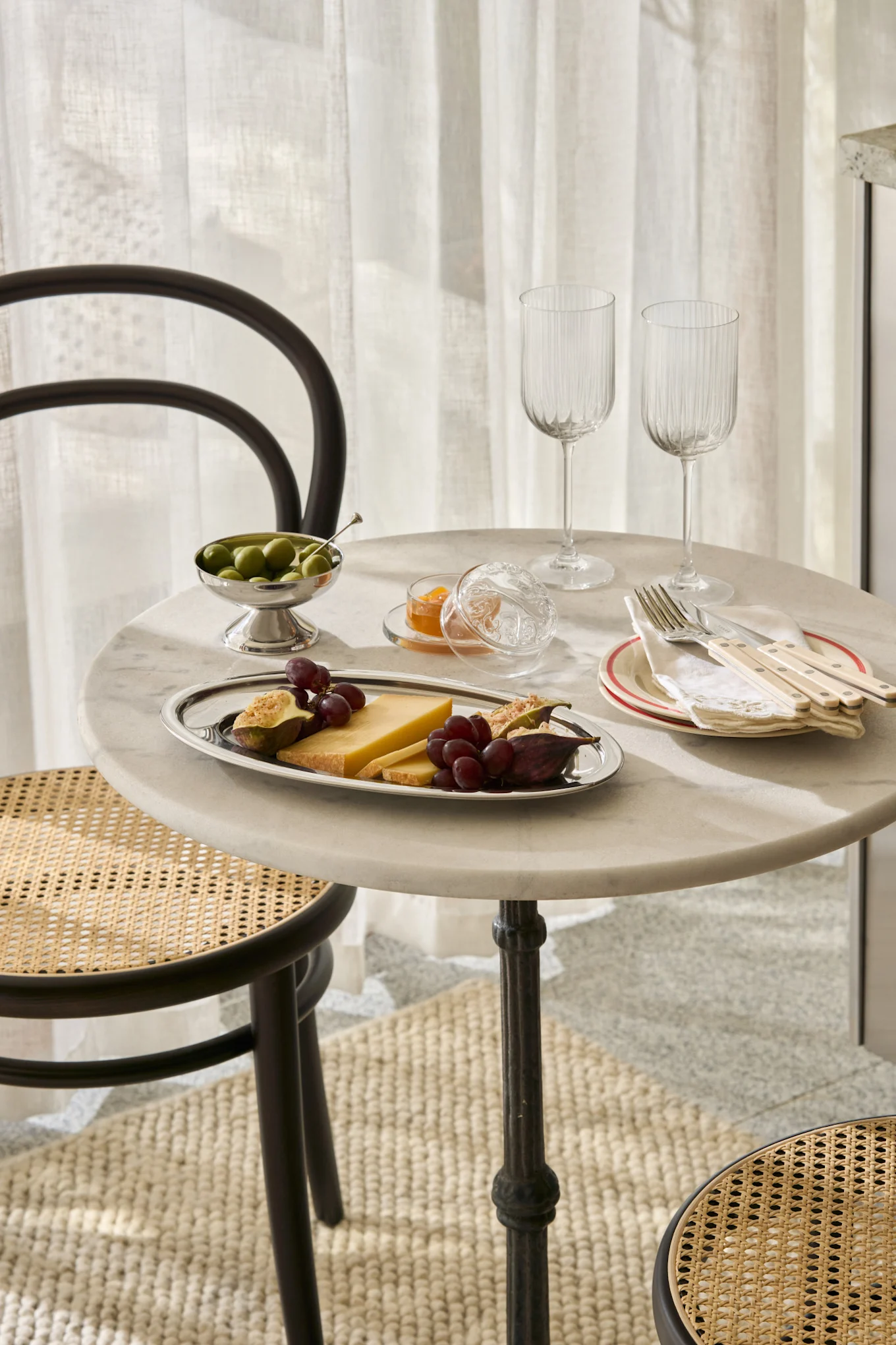 Una mesa de mármol puesta con queso, higos, uvas, aceitunas, mermelada, copas y cubiertos, con sillas de mimbre.