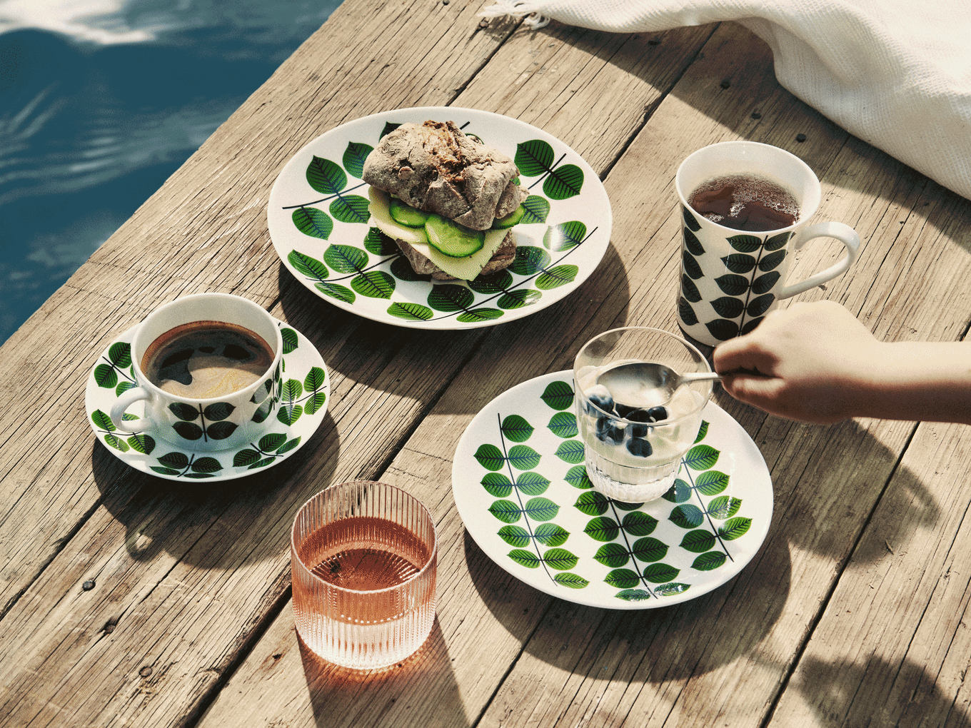 Desayuno en muelle de madera: platos con hojas, sándwich, café, té, yogur con arándanos y bebida rosa.