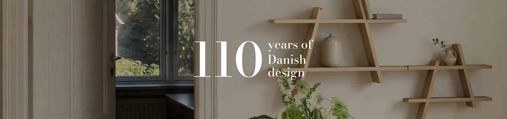 Texto '110 years of Danish design' sobre una toma interior de una habitación con una estantería geométrica de madera, un jarrón y flores.