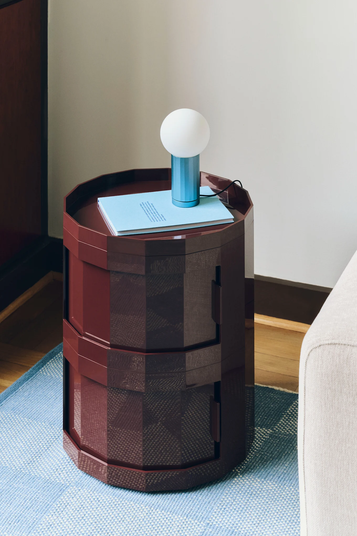Mesa auxiliar hexagonal rojo oscuro con un libro azul claro y una pequeña lámpara azul y blanca encima, sobre una alfombra azul.