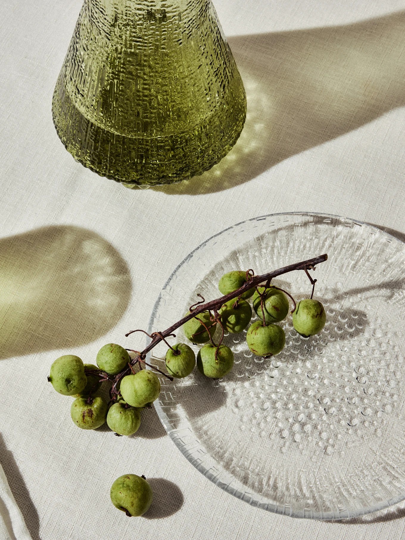 Jarrón de cristal verde texturizado y un plato transparente con pequeñas frutas verdes en una rama.