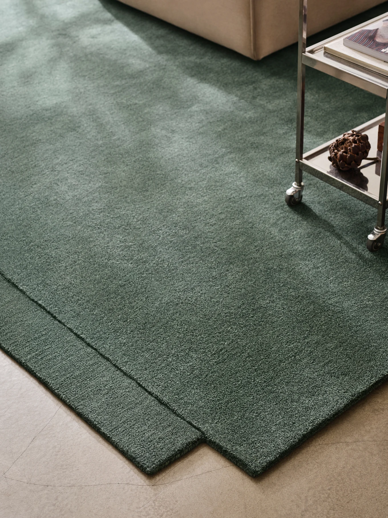 Alfombra verde oscuro de pelo largo en suelo de hormigón, con sofá beige y carrito metálico.