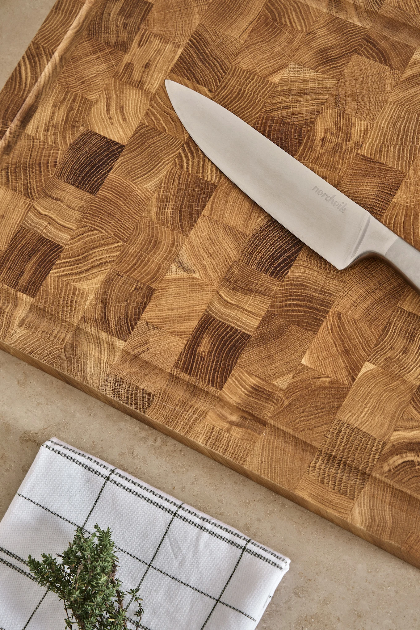 Cuchillo de chef Nordwik sobre tabla de cortar de madera con veta de testa, paño a cuadros y tomillo fresco.