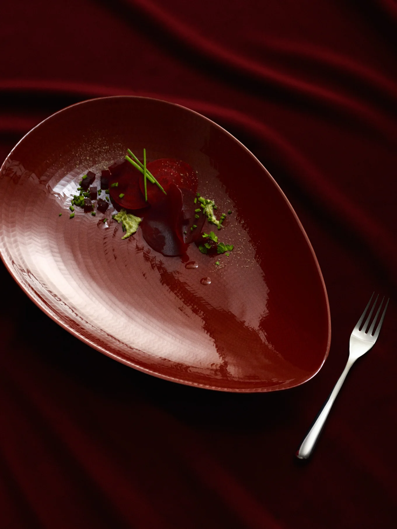 Un plato rojo oscuro en forma de pera con un plato de remolacha, adornado con hierbas verdes y cubos oscuros, junto a un tenedor plateado sobre una tela roja oscura drapeada.