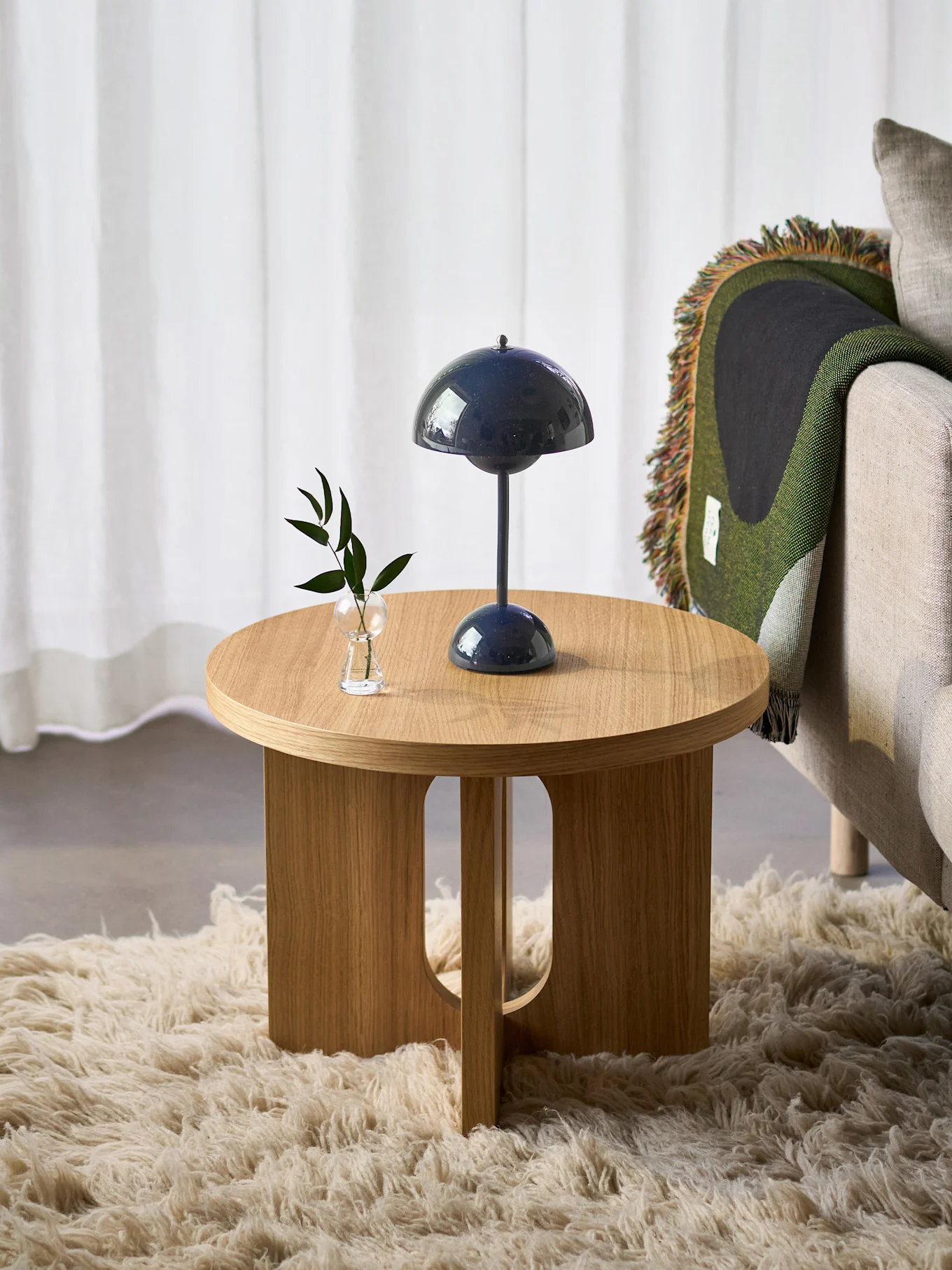 Mesa auxiliar redonda de madera con lámpara azul y planta, junto a sillón con manta verde, sobre alfombra peluda.