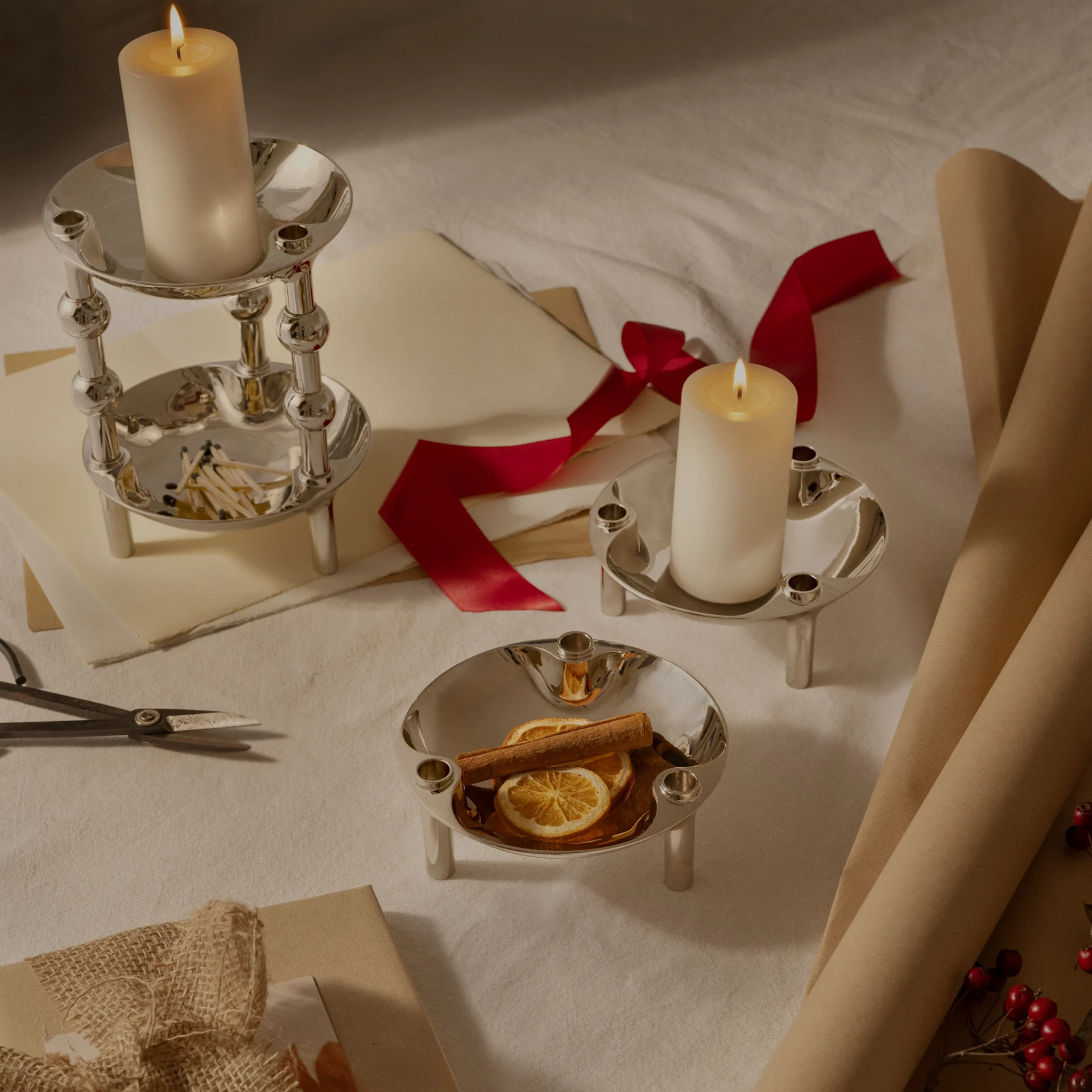 Tres candelabros de plata, dos con velas encendidas, uno con naranjas secas y canela, dispuestos con papel de regalo y cinta.