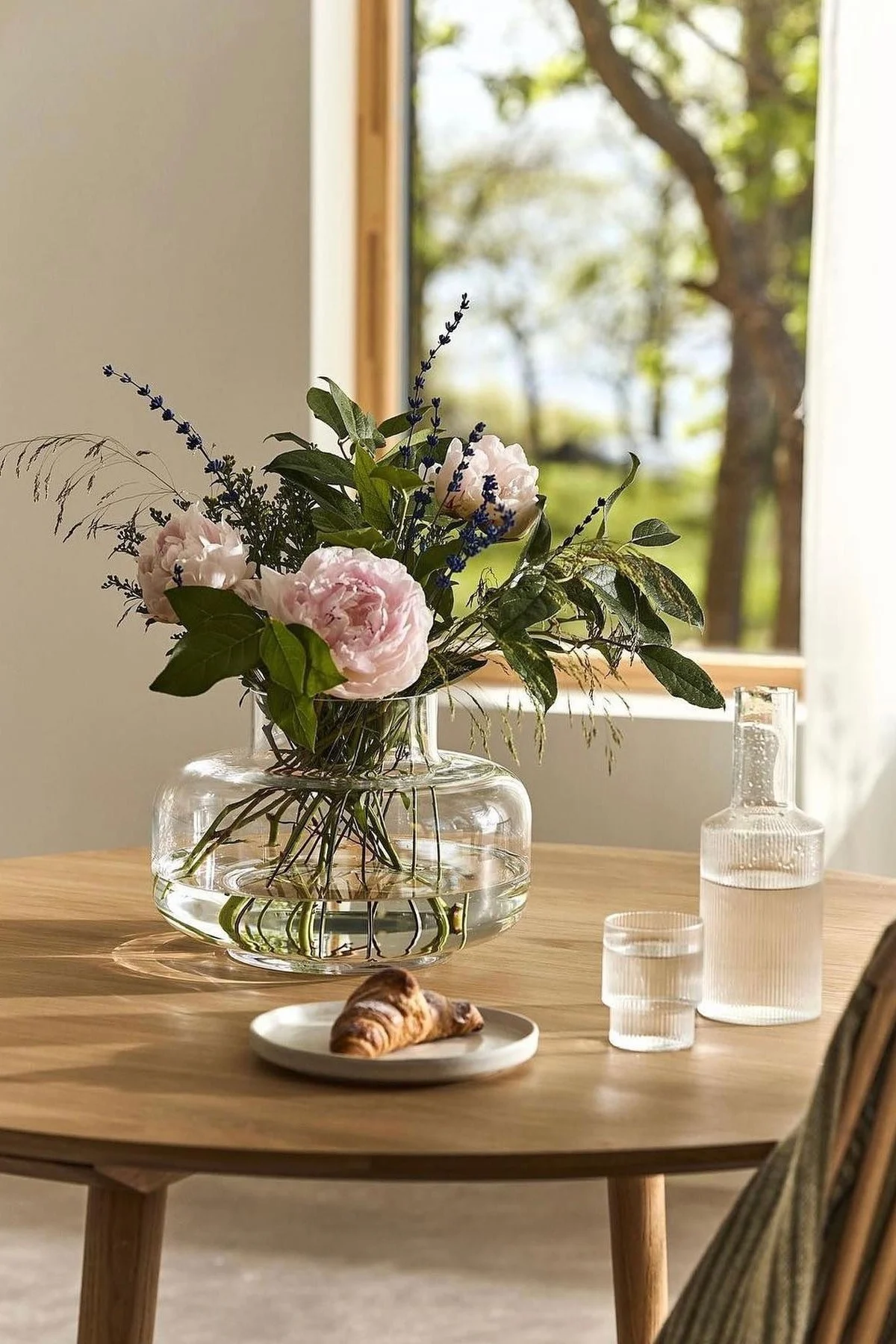 En una mesa puesta con una jarra y un croissant se encuentra el jarrón Urna de Marimekko con peonías.