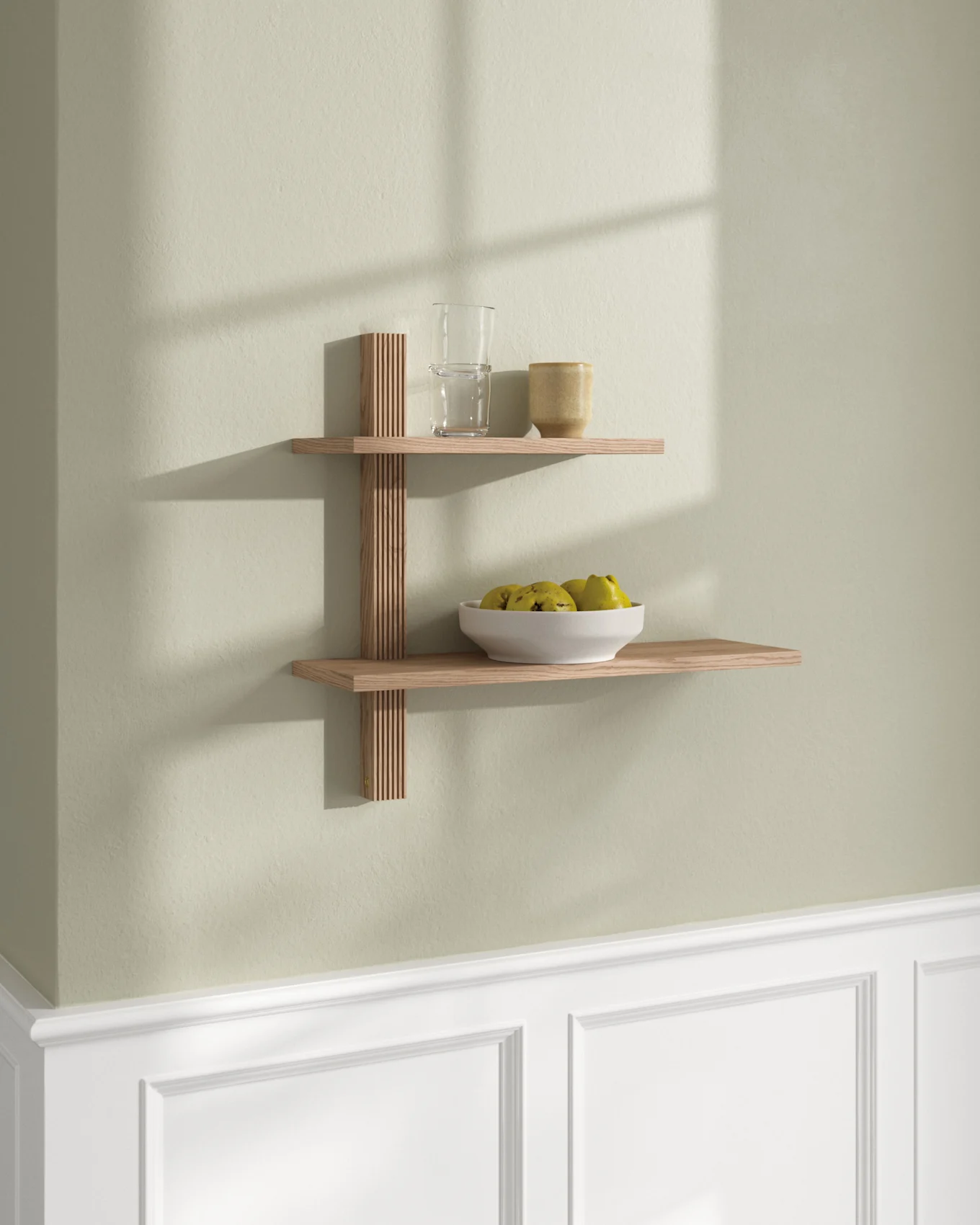 Dos estantes flotantes de madera clara en una pared verde claro. El estante superior tiene un vaso y una taza; el inferior, un cuenco de fruta.
