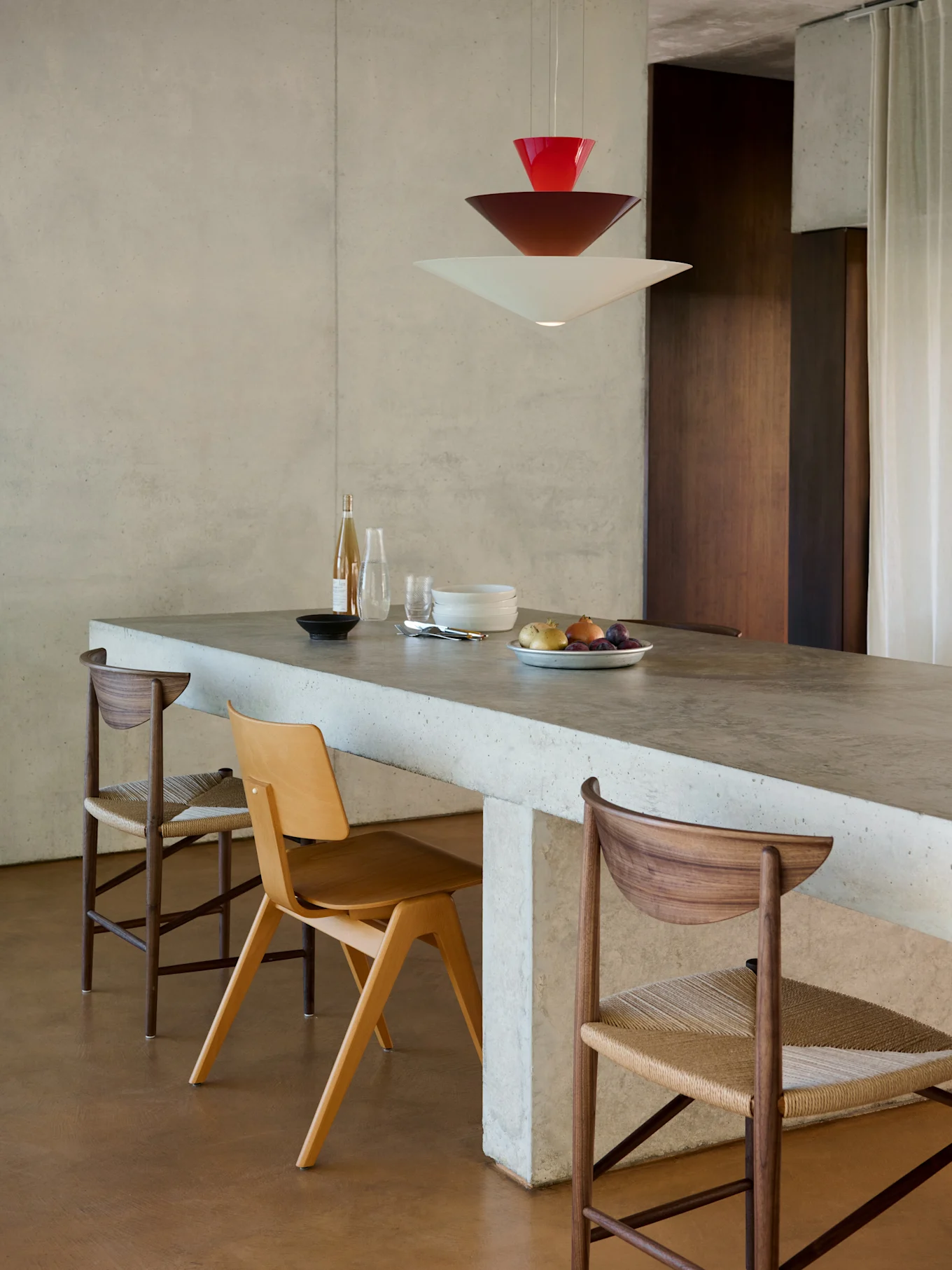 Comedor moderno con isla de hormigón, tres sillas y lámpara colgante apilada roja, marrón y blanca.
