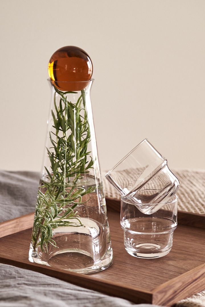 Aquí puedes ver la Jarra Fia con tapa ámbar y una ramita de romero con los Vasos NM&, todos de Design House Stockholm.