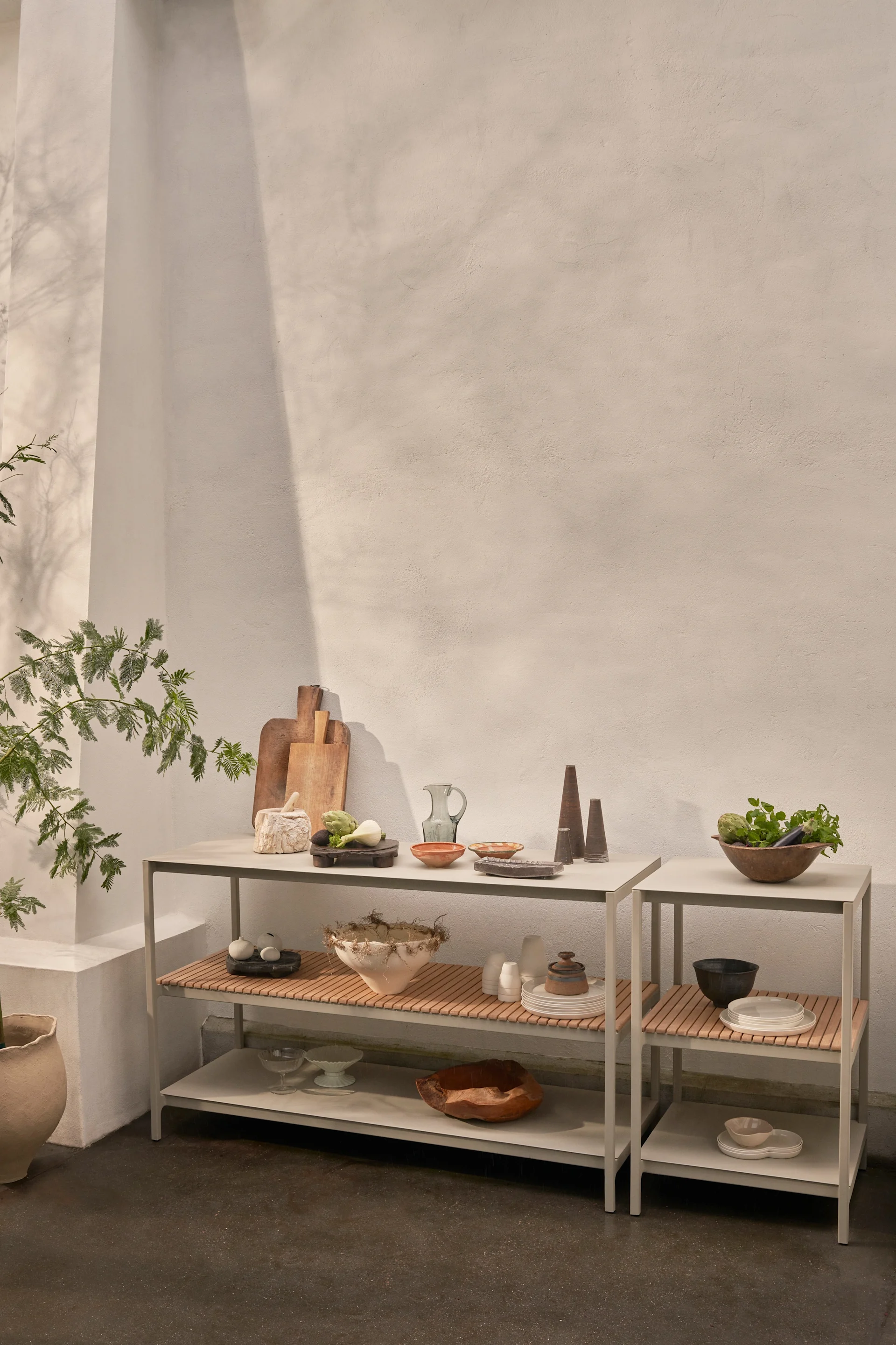 Dos estanterías grises modernas con tapas de lamas de madera muestran menaje de cocina, cuencos de cerámica, verduras frescas y objetos decorativos contra una pared clara.