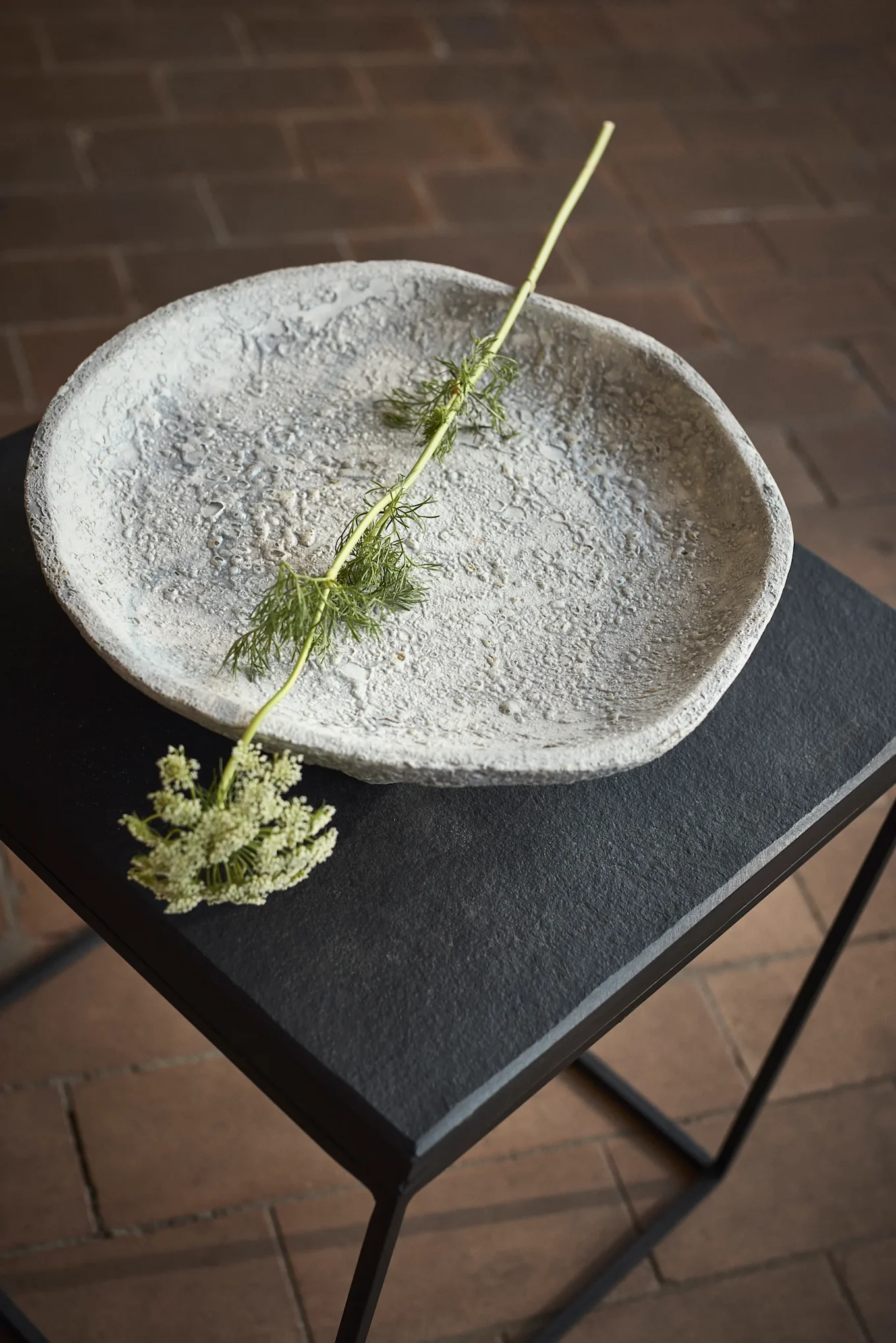 Cuenco gris texturizado sobre una mesa negra con un tallo verde y pequeñas flores blancas.