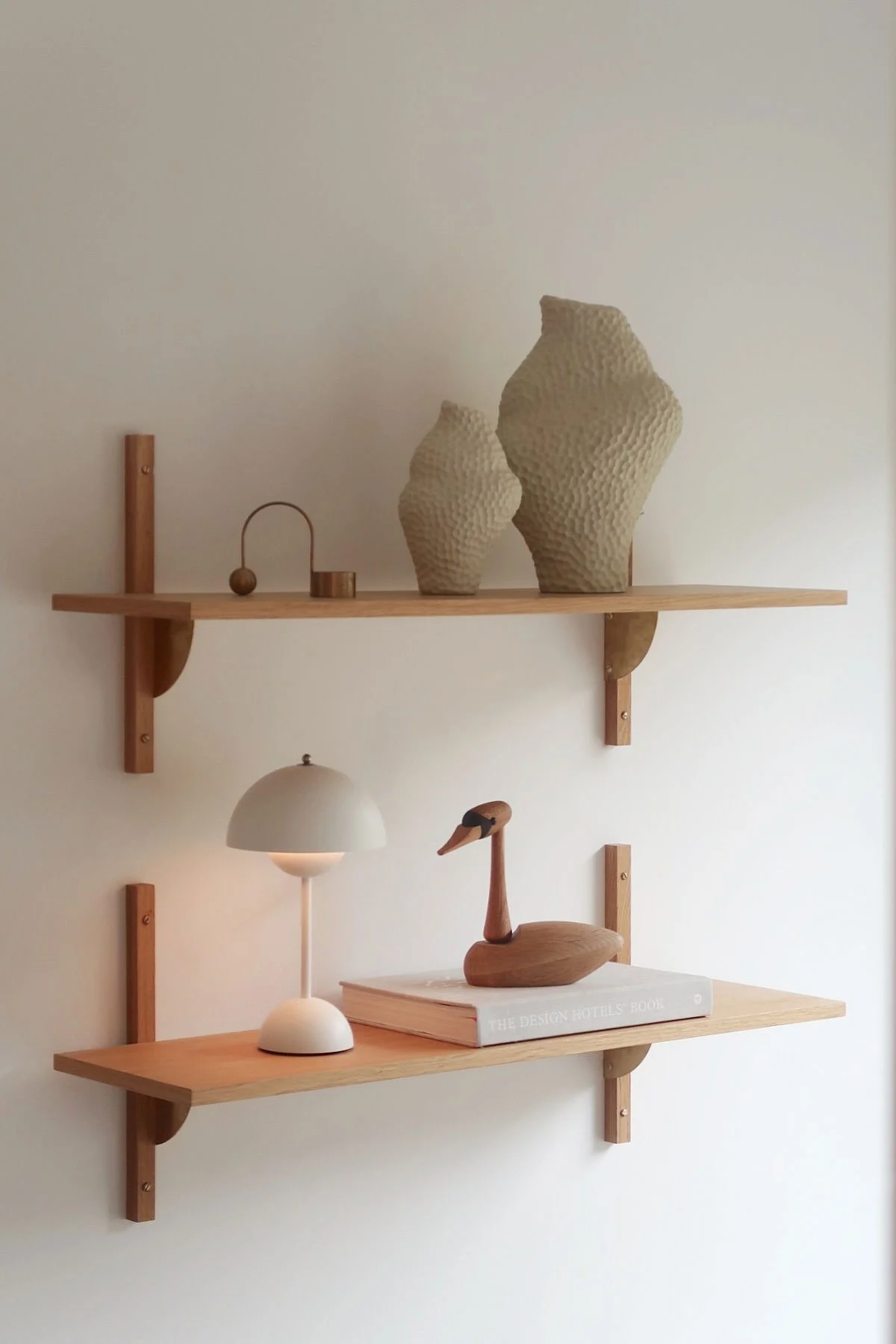 Dos estantes de pared de madera clara con decoración. El estante superior contiene dos jarrones texturizados y objetos de metal. El estante inferior tiene una lámpara blanca, un cisne de madera y un libro.
