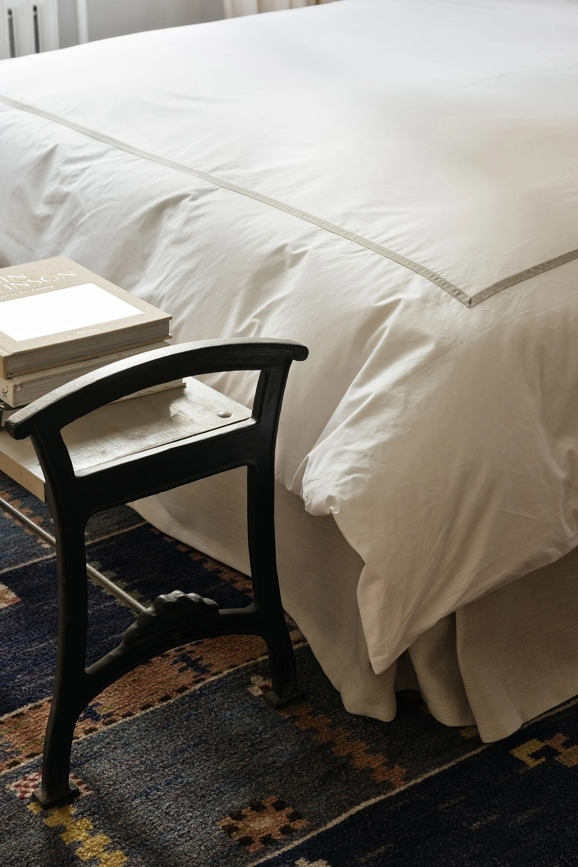 Una cama con ropa de cama blanca y una raya gris junto a un taburete oscuro con libros sobre una alfombra azul estampada.