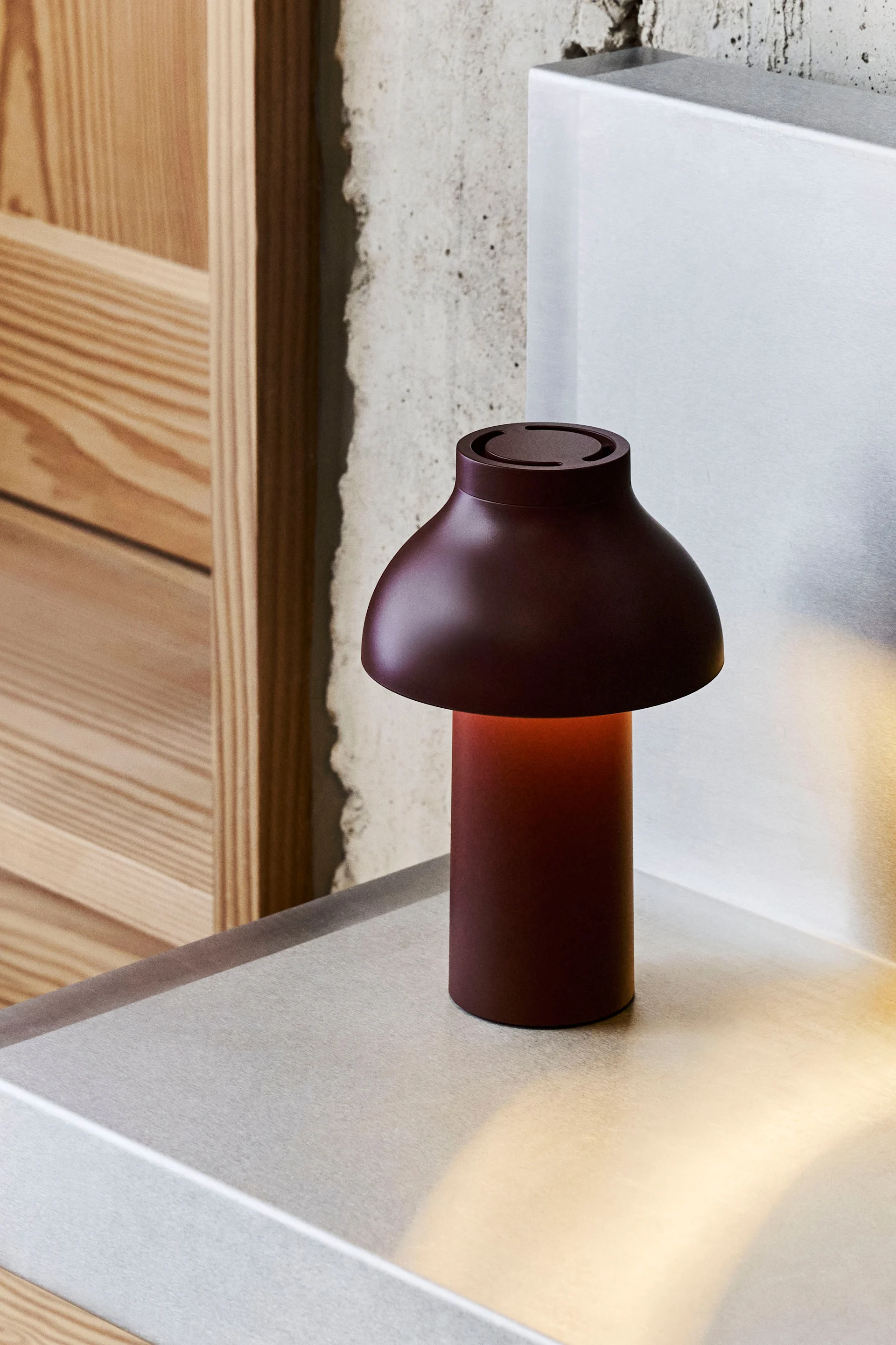Una lámpara de mesa en forma de hongo rojo oscuro con un brillo cálido sobre una superficie metálica, con hormigón y madera de fondo.