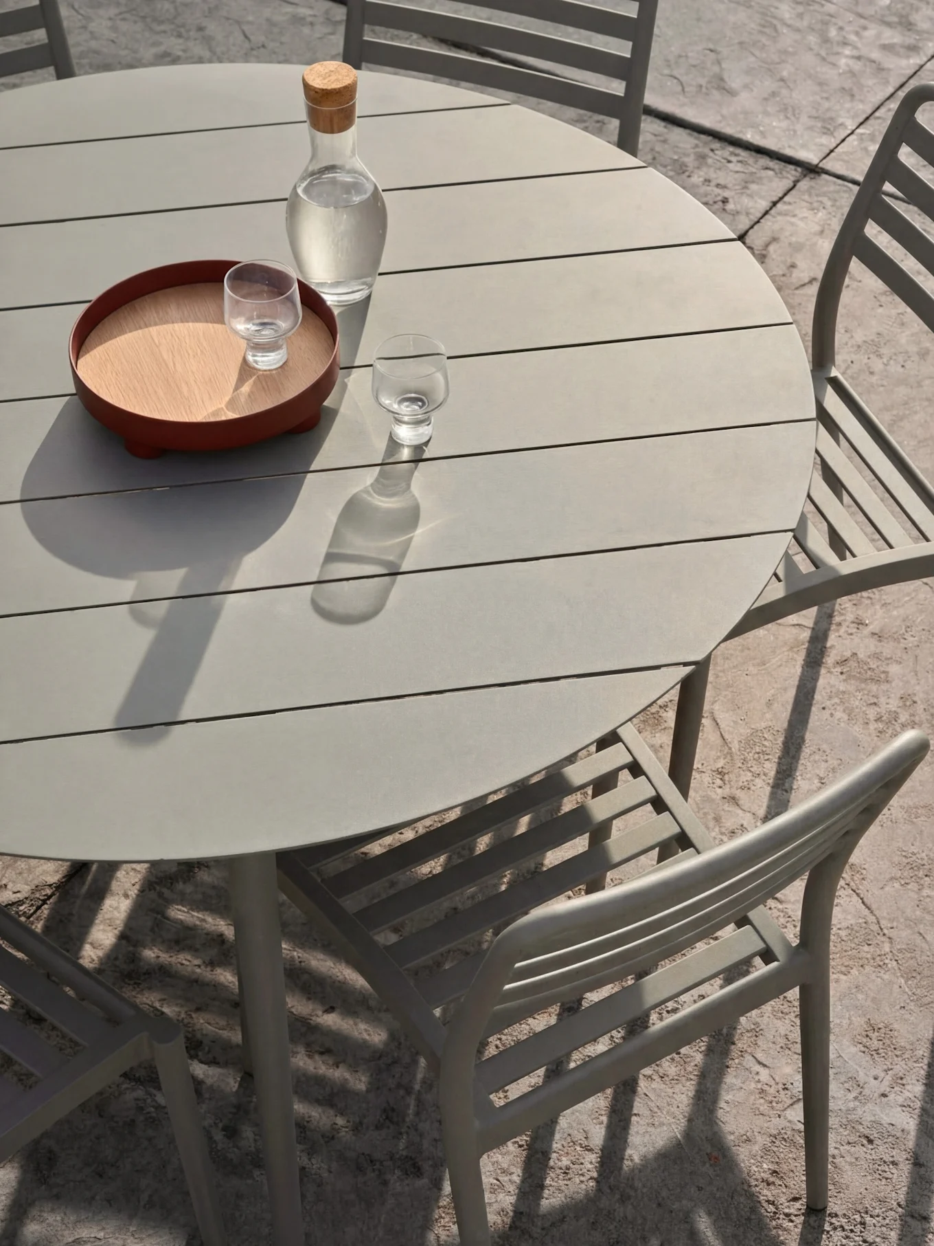 Un juego de comedor exterior gris claro con una mesa redonda de listones y sillas. Sobre la mesa, una jarra de agua y vasos en una bandeja.