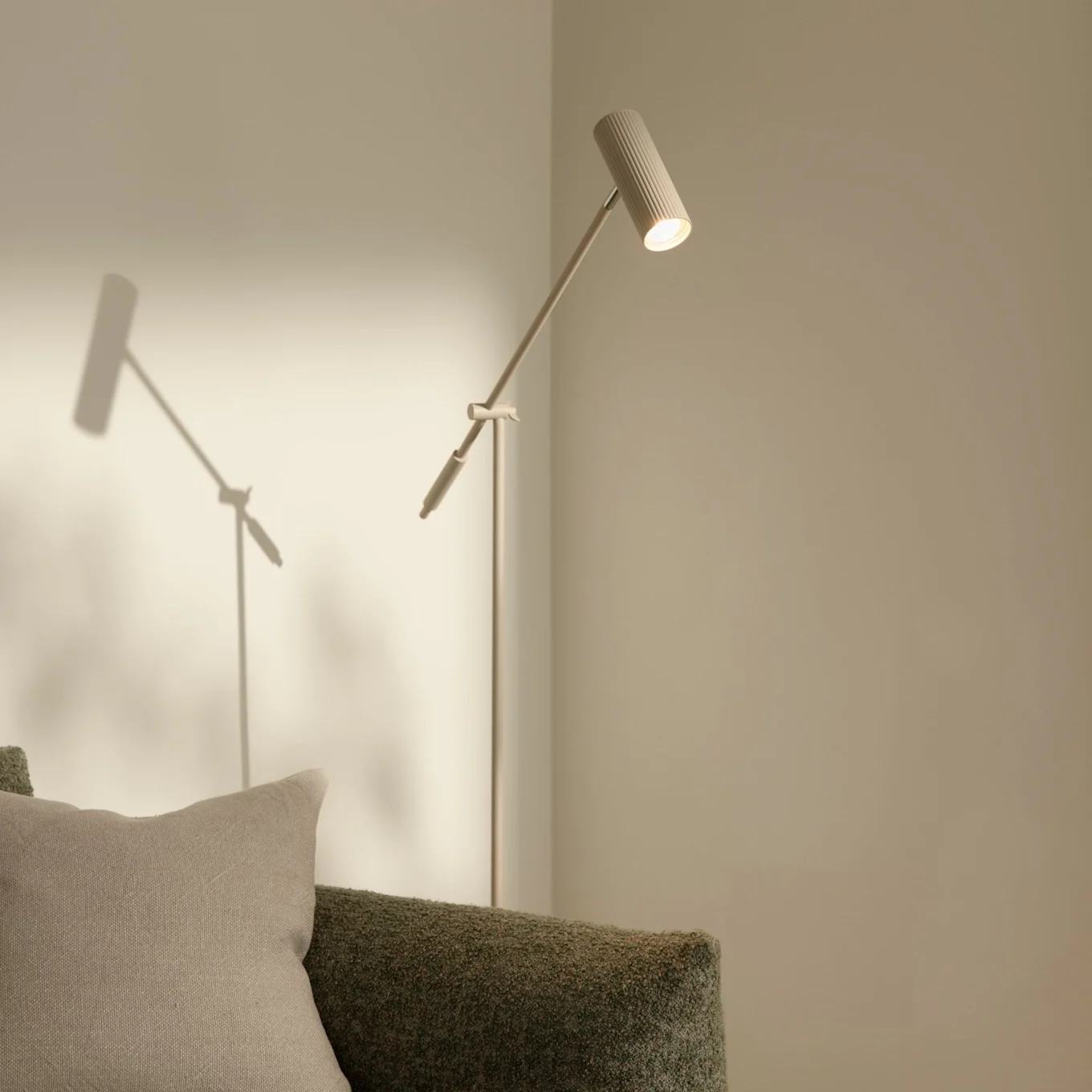 Una lámpara de pie moderna y esbelta con cabezal estriado que ilumina una pared clara, junto a un sofá verde texturizado con un cojín beige.