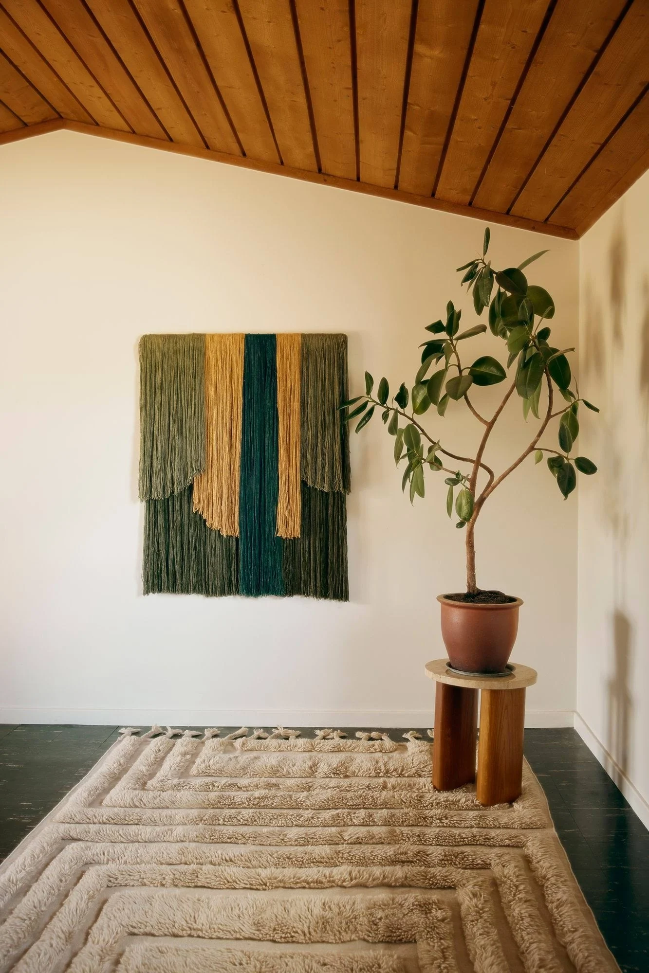 Una habitación con un tapiz de pared de hilo texturizado verde, dorado y verde azulado, una planta en maceta y una alfombra beige peluda con patrones geométricos.