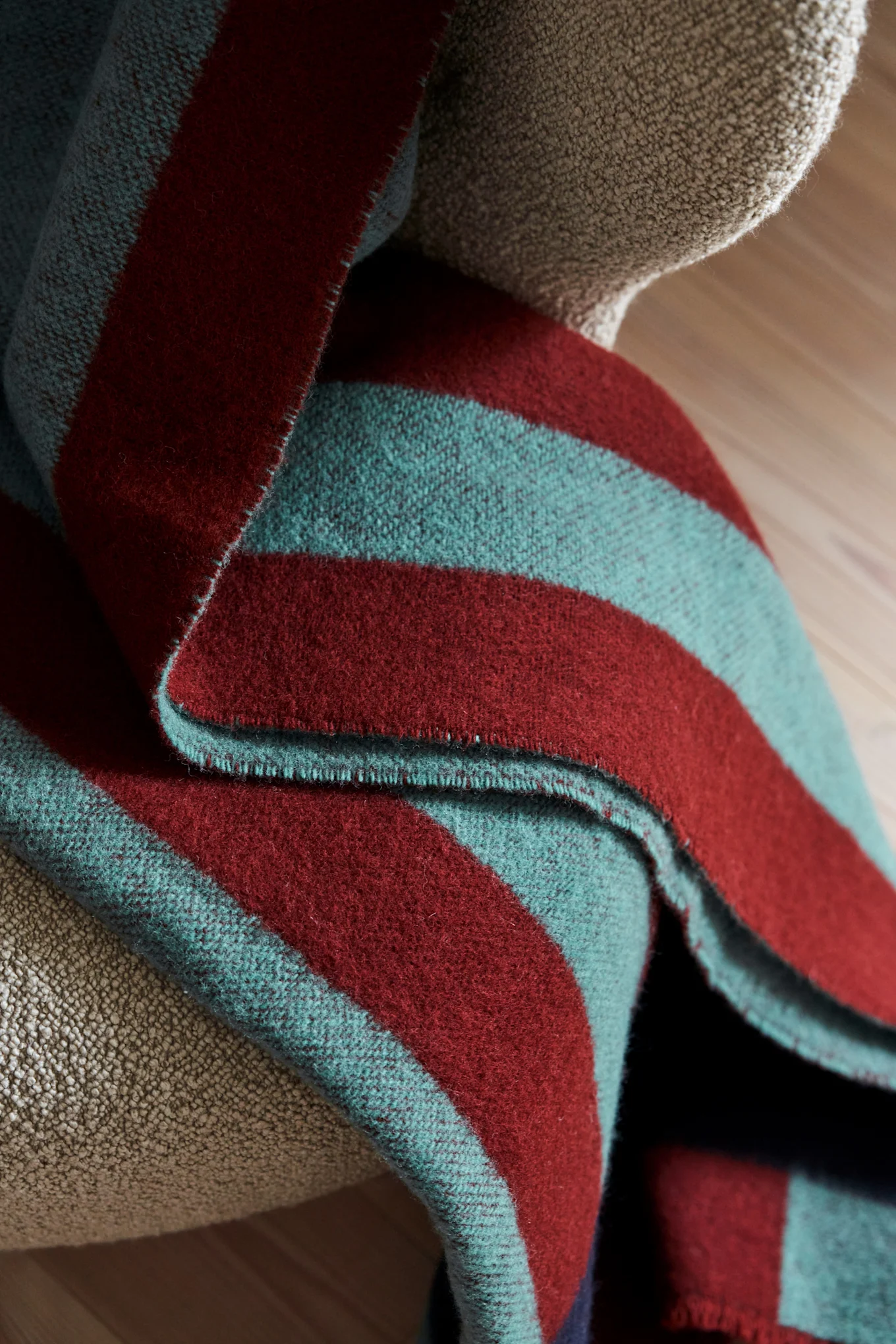 Manta a rayas doblada con bandas rojas oscuras y verde azulado sobre un sillón texturizado de color marrón claro.