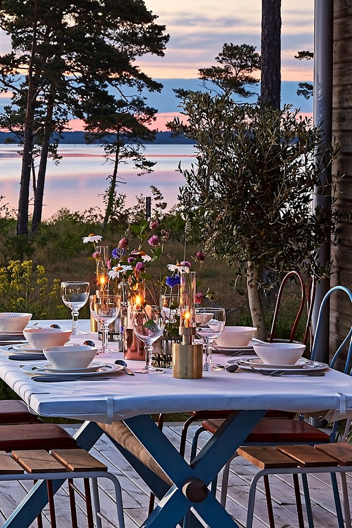 Farolillos y portavelas para exteriores: aquí puedes ver una mesa de madera en una terraza con el mar de fondo durante la puesta de sol. La mesa está puesta y decorada con la lámpara de aceite Patina de Klong, entre otras cosas.