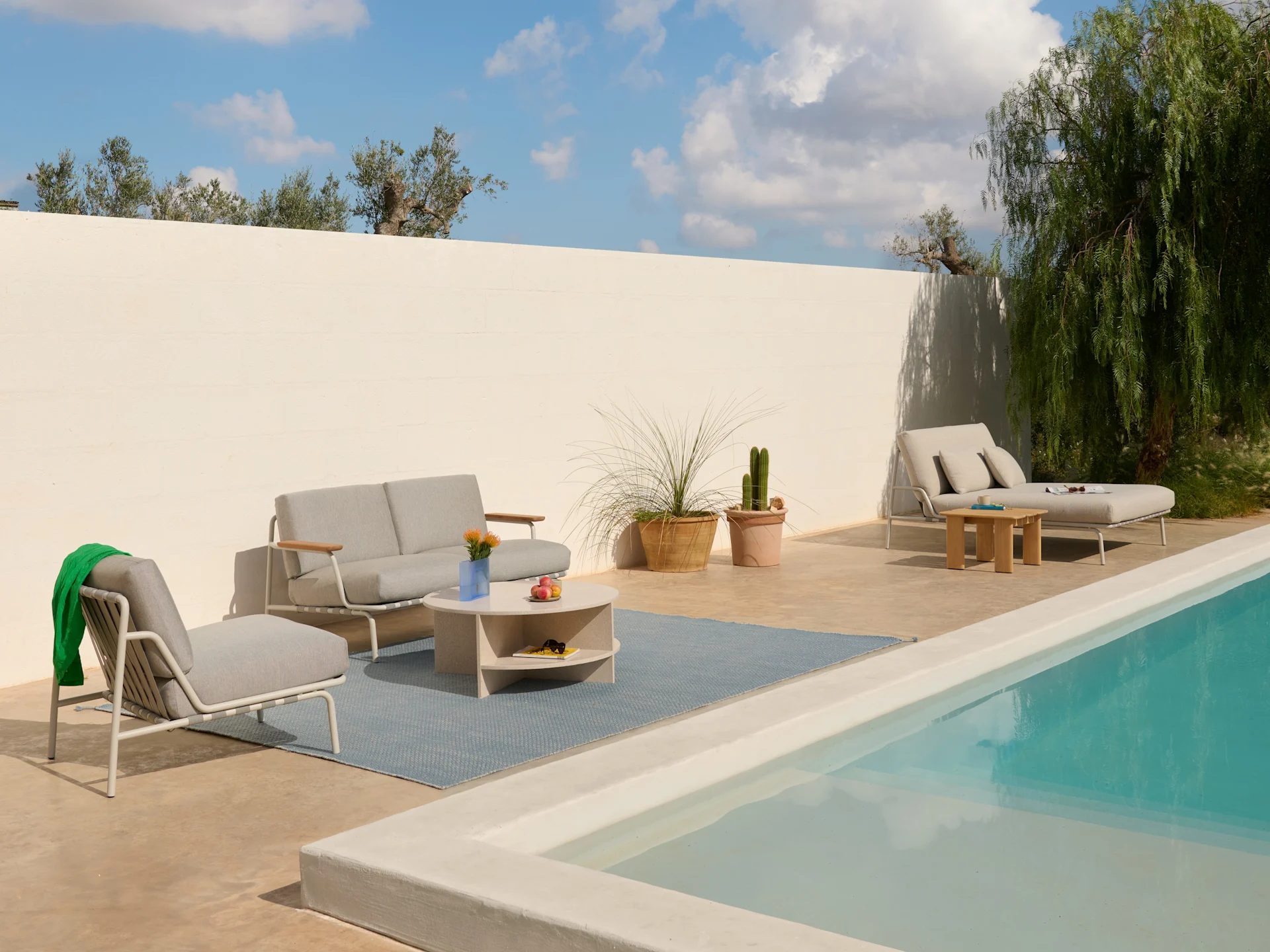 Patio exterior con sofá gris claro, sillones, mesa de centro sobre una alfombra azul y una tumbona junto a una piscina.