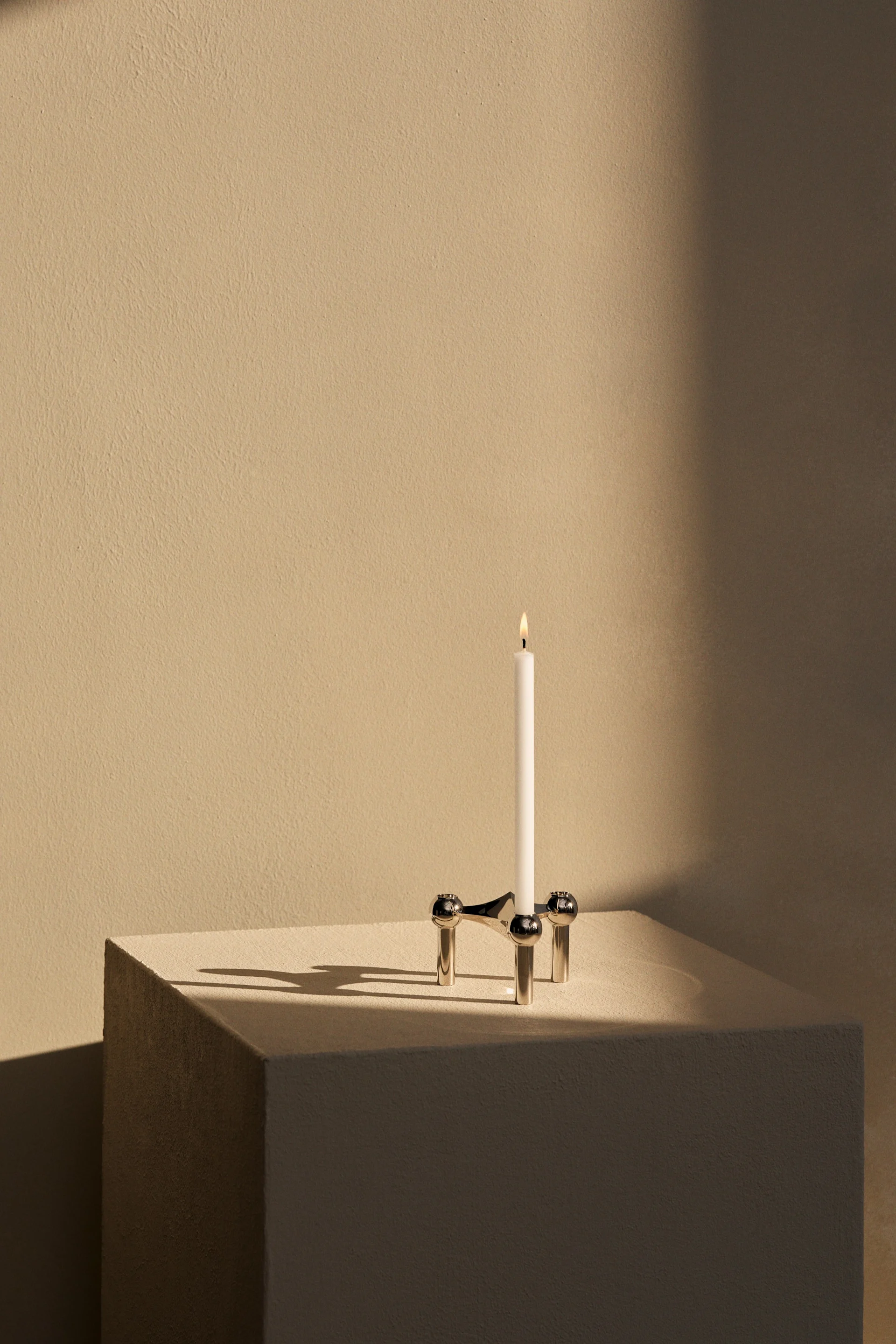 Vela blanca encendida en candelabro plateado de tres patas con detalles esféricos, sombras en superficie beige clara texturizada.