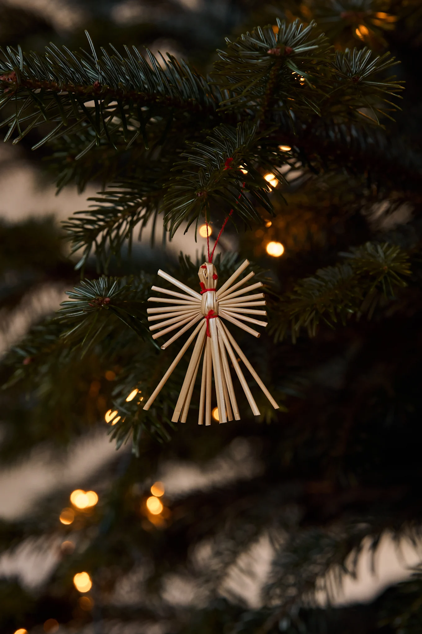 Adorno de ángel de paja colgando de una rama de árbol de Navidad con luces borrosas al fondo.