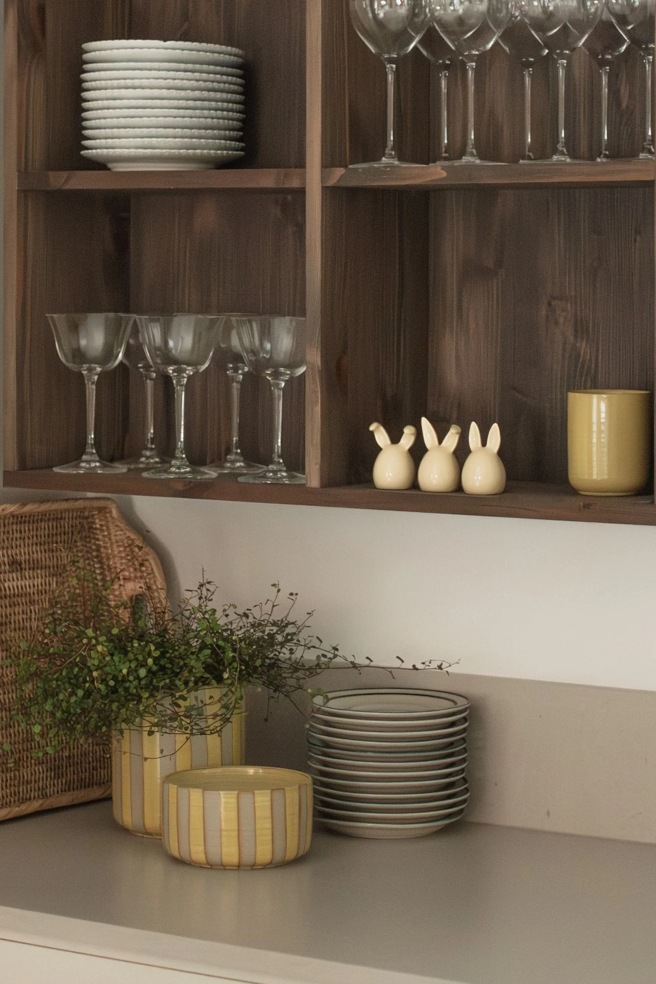 Estantes de madera con platos blancos apilados, vasos variados, tres conejitos de cerámica y una taza amarilla. En el mostrador, una cesta tejida, plantas en macetas a rayas y más platos.