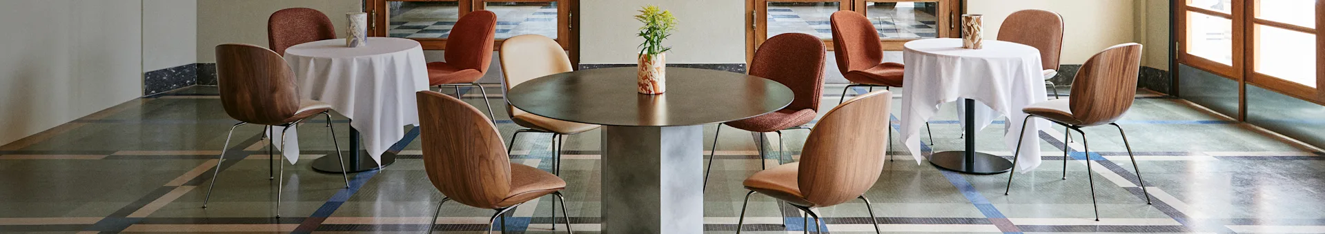 Interior elegante de cafetería con una mesa central grande, dos mesas más pequeñas con manteles blancos y varias sillas sobre un suelo estampado.