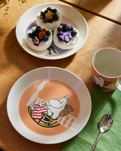 Plato, taza y cuchara de Moomintroll junto a un plato de merengues con arándanos y flores en una mesa de madera.