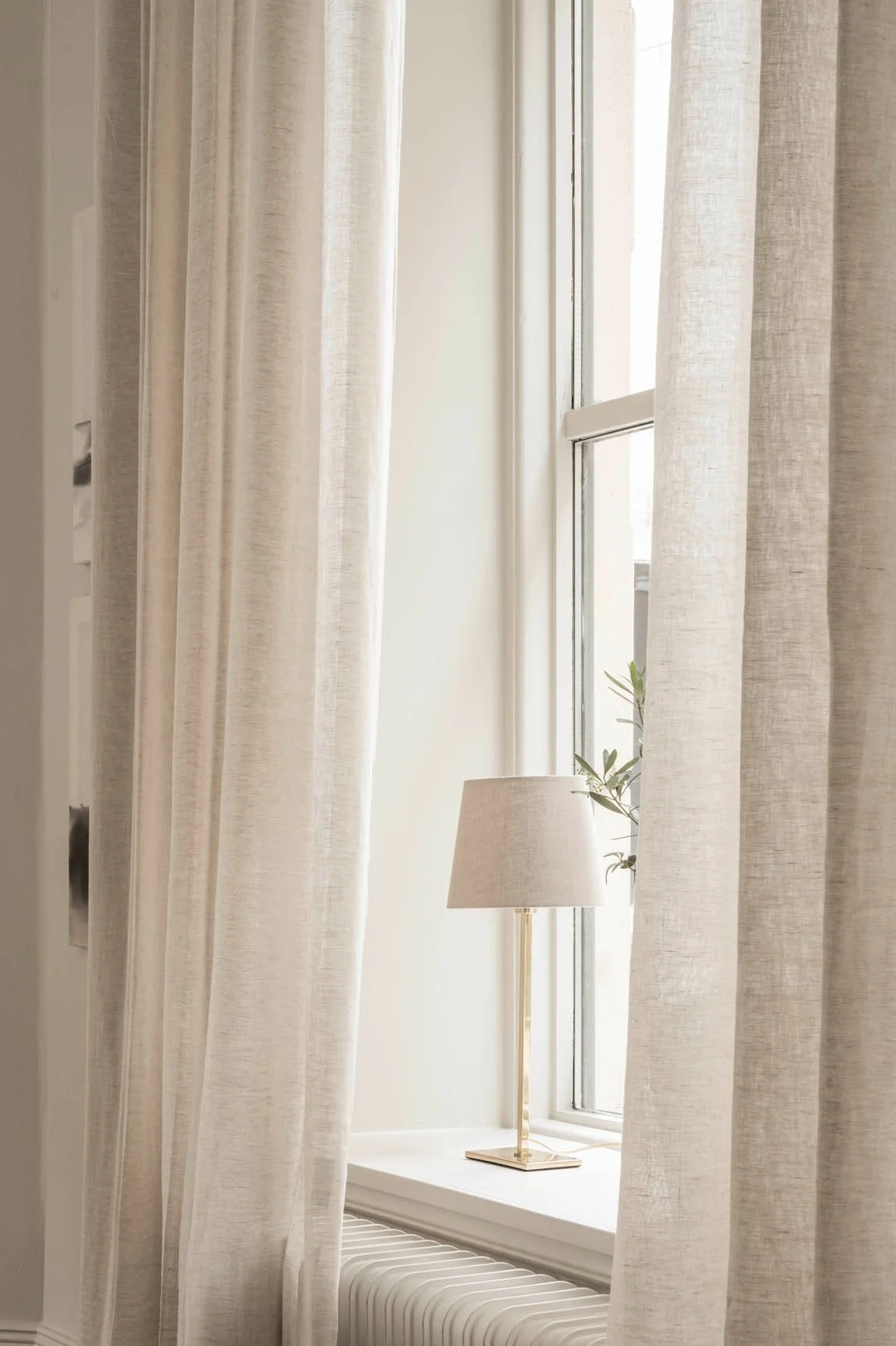 Una lámpara de sobremesa de Watt & Veke se encuentra en el alféizar de una ventana, rodeada de cortinas claras.