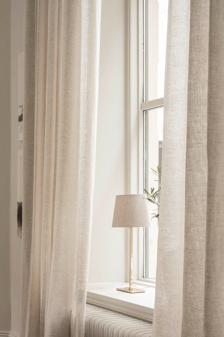 Una lámpara de sobremesa de Watt & Veke se encuentra en el alféizar de una ventana, rodeada de cortinas claras.