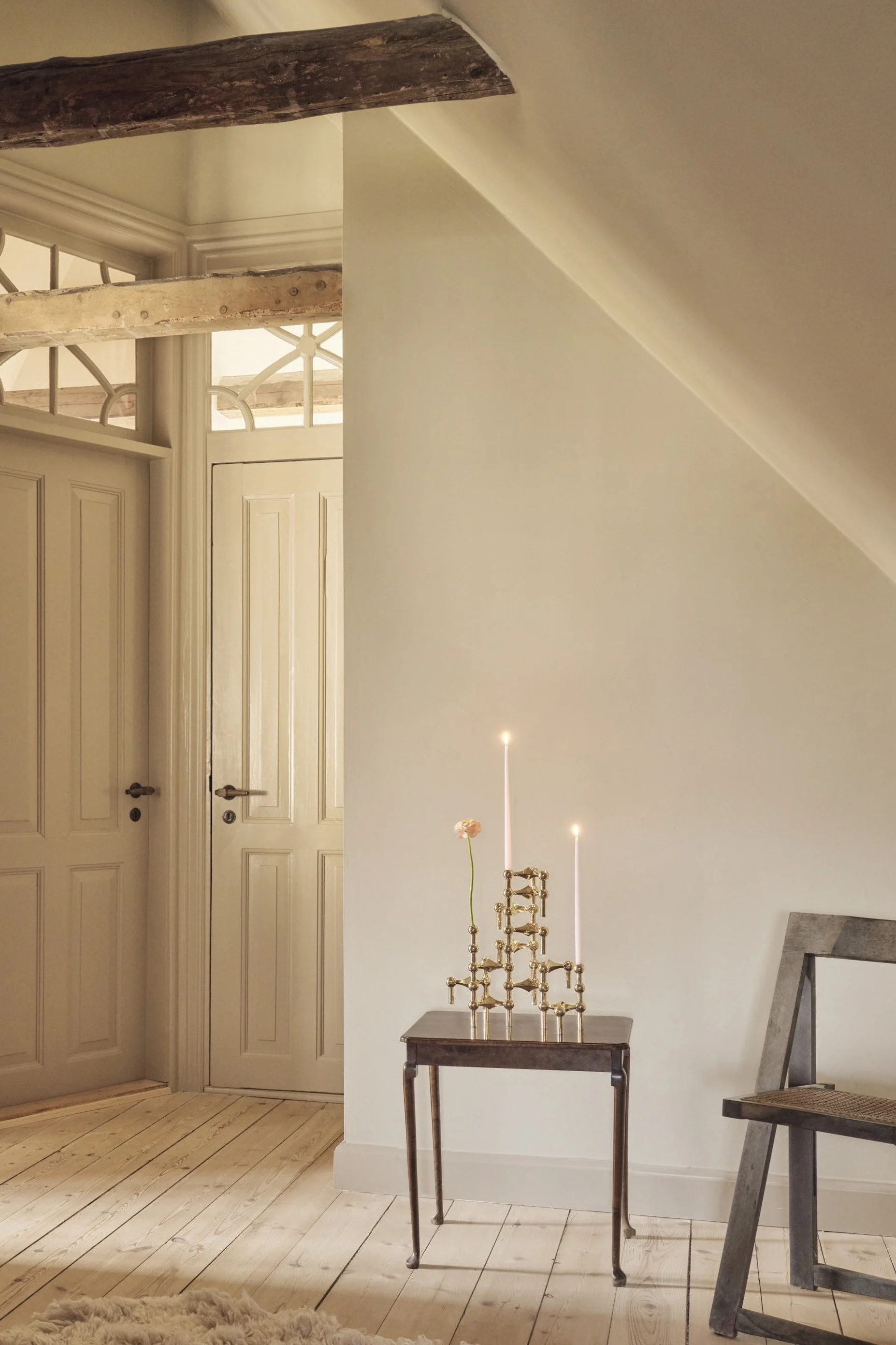 Una habitación abuhardillada con paredes claras y suelos de madera. Dos puertas con paneles y tragaluces ornamentados, una viga expuesta, una mesa auxiliar con velas encendidas y una flor, una silla y una alfombra mullida.