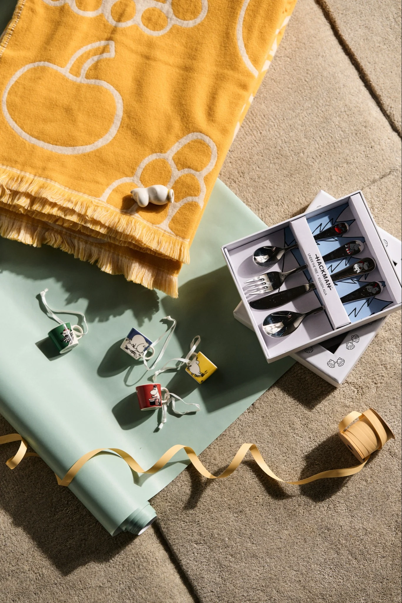 Ideas creativas de regalos para una fiesta baby shower, aquí puedes ver una colección de posibles regalos para un baby shower. Hay mini tazas, figuritas de Mumin, una manta amarilla y un juego de cubiertos infantiles.