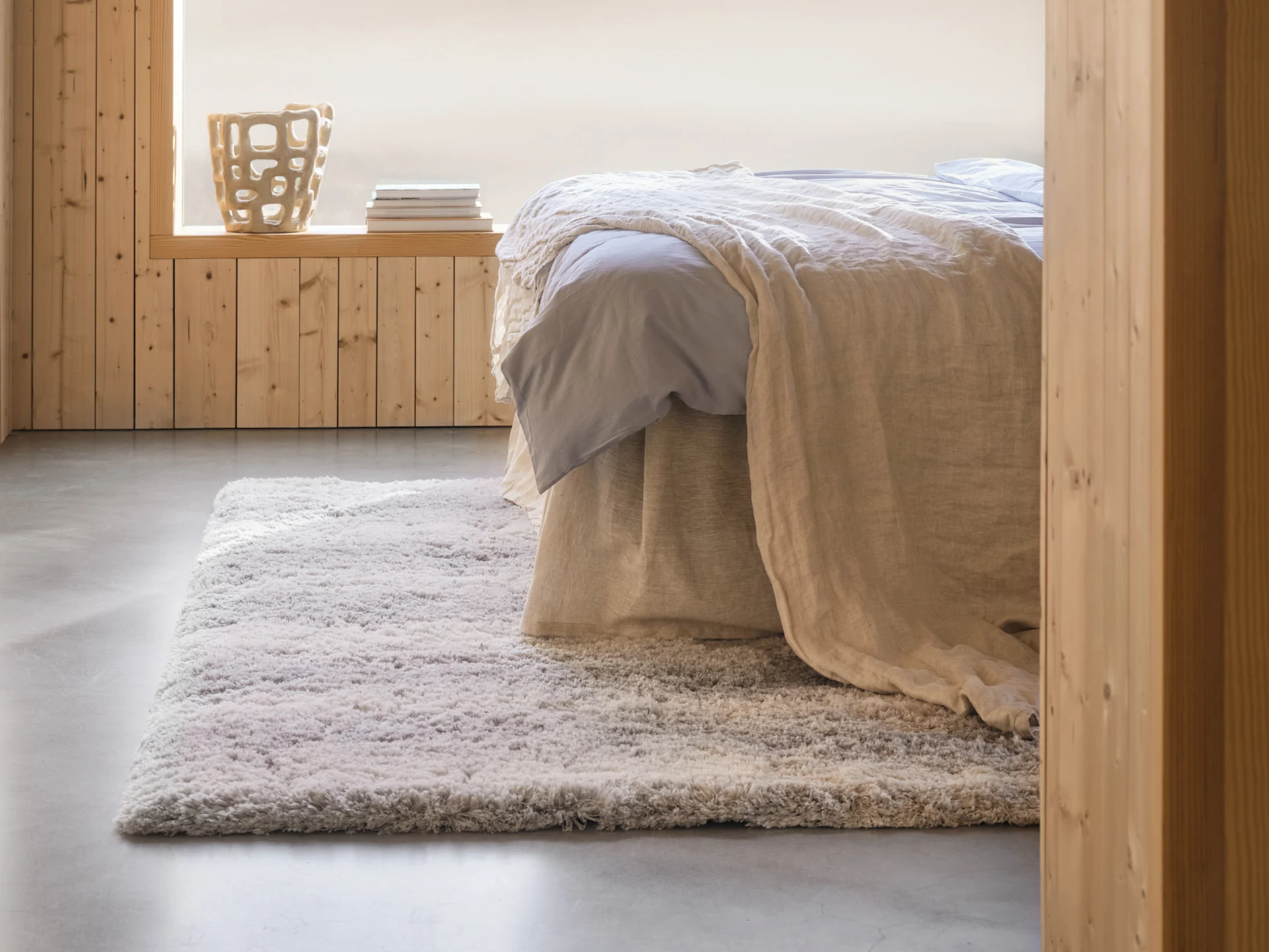 Acogedor dormitorio con cama, sábanas azul claro, manta beige, alfombra peluda, paredes de madera y un jarrón decorativo.