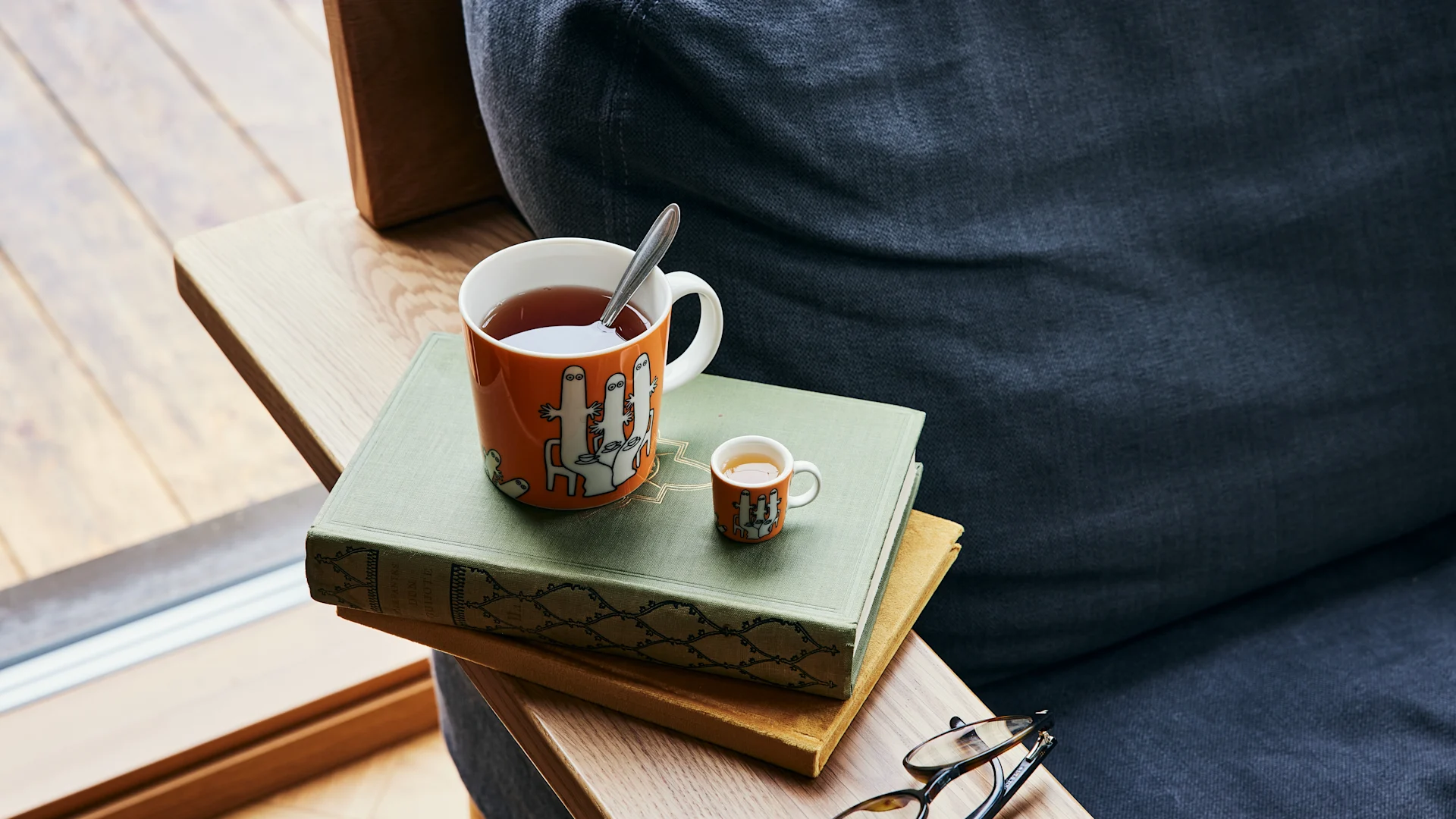 La taza de color naranja del personaje Mumin Hatifnates, con té está junto a un sillón sobre una pila de libros.