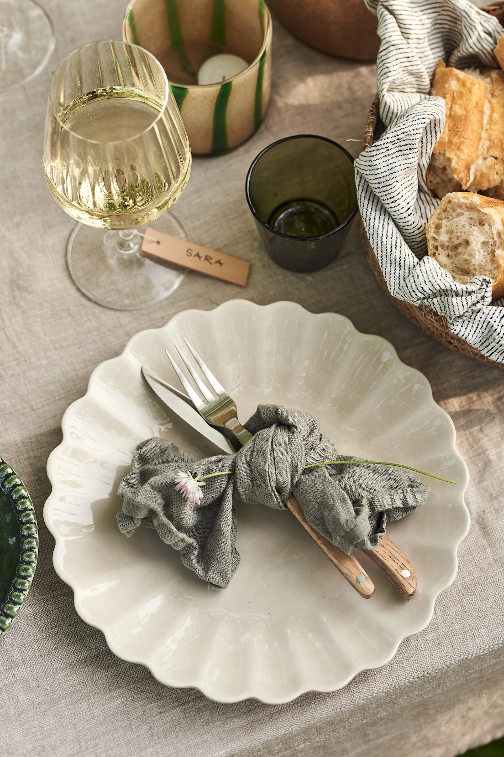 Conjunto de mesa de comedor para exterior con un plato blanco festoneado, cubiertos atados con una servilleta gris, una copa de vino y una cesta de pan.
