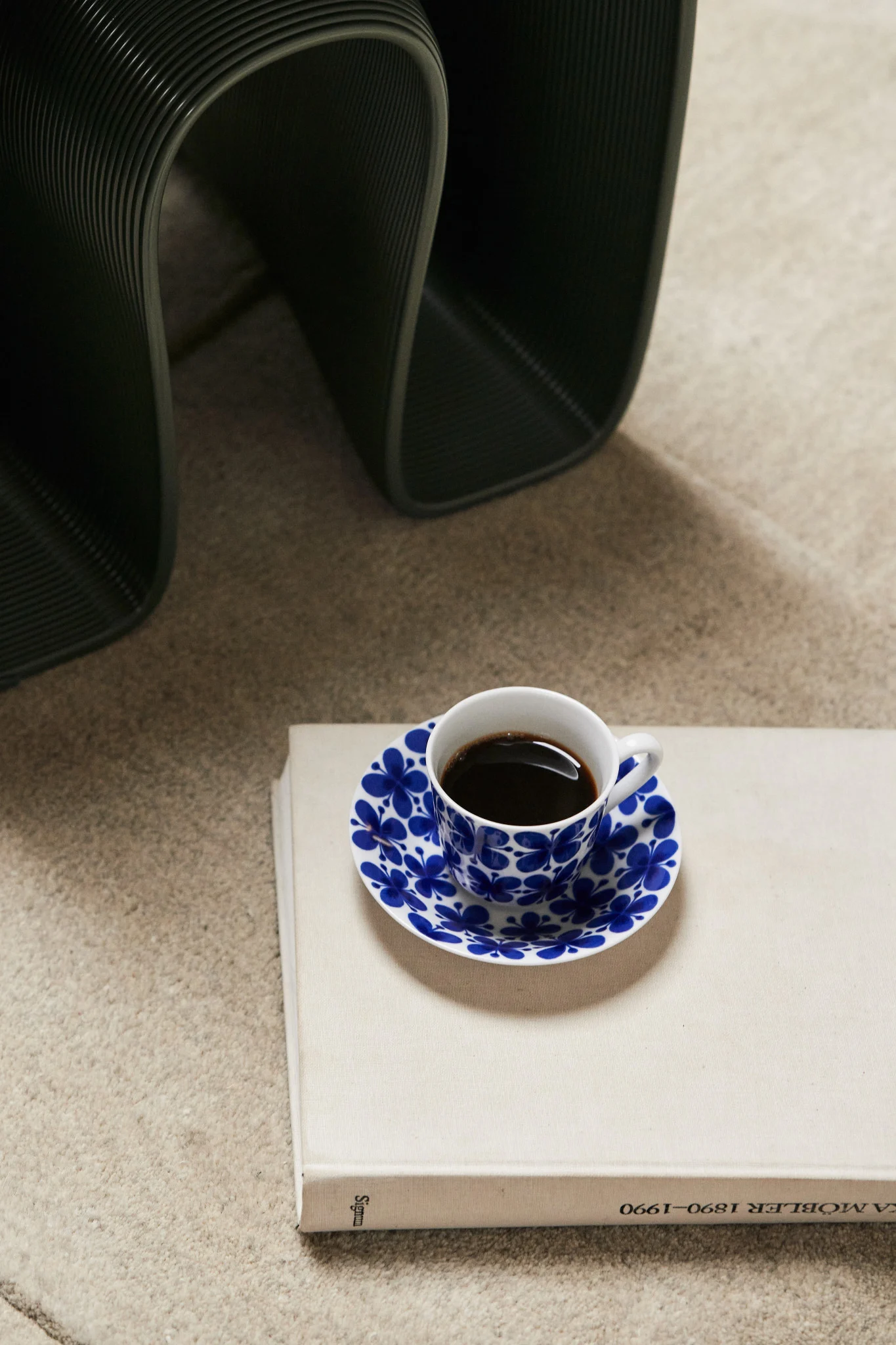 Taza de café con platillo floral azul sobre un libro claro, objeto oscuro acanalado al fondo.