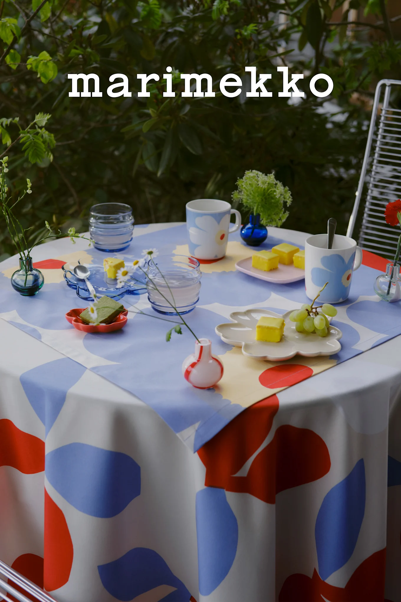 Logotipo de Marimekko. Una mesa exterior puesta con un mantel de patrón abstracto azul, rojo y blanco, tazas florales, pasteles amarillos, uvas y pequeños jarrones.