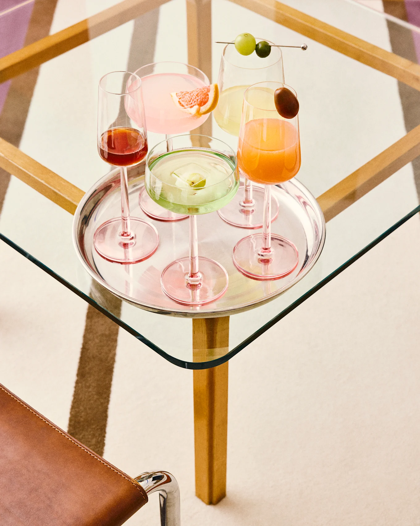 Cinco cócteles coloridos en una bandeja de plata: rosa con pomelo, verde con hielo de frutas, amarillo con uvas, naranja con una guarnición y rojo oscuro, todos sobre una mesa de cristal.