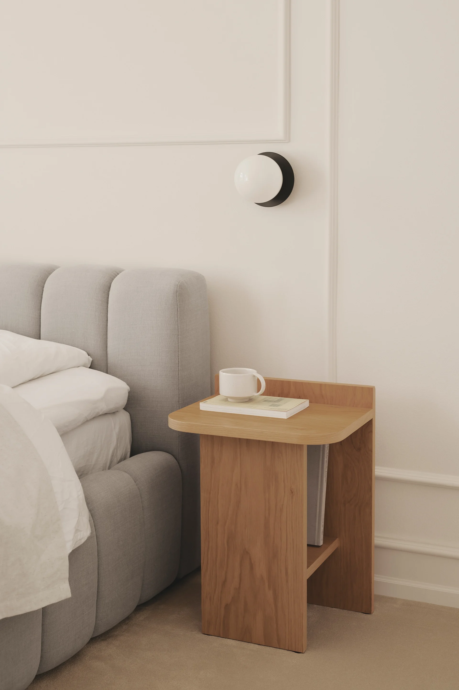 Un dormitorio acogedor con una cama tapizada gris junto a una mesita de noche de madera con una taza y un libro, y una lámpara de pared circular.