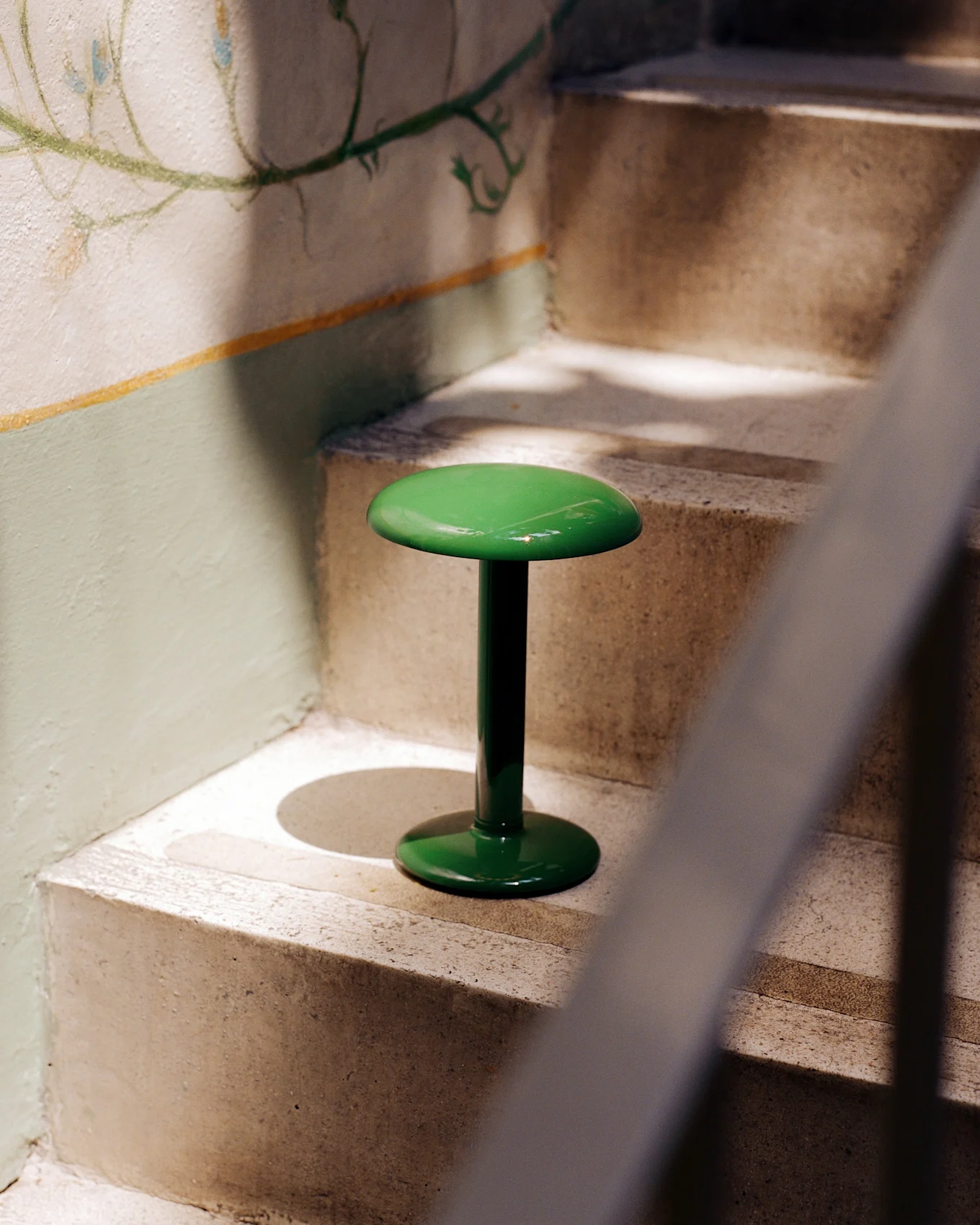 Un taburete verde brillante y brillante con tapa y base redondas se encuentra en una escalera de hormigón junto a una pared con un mural floral descolorido.
