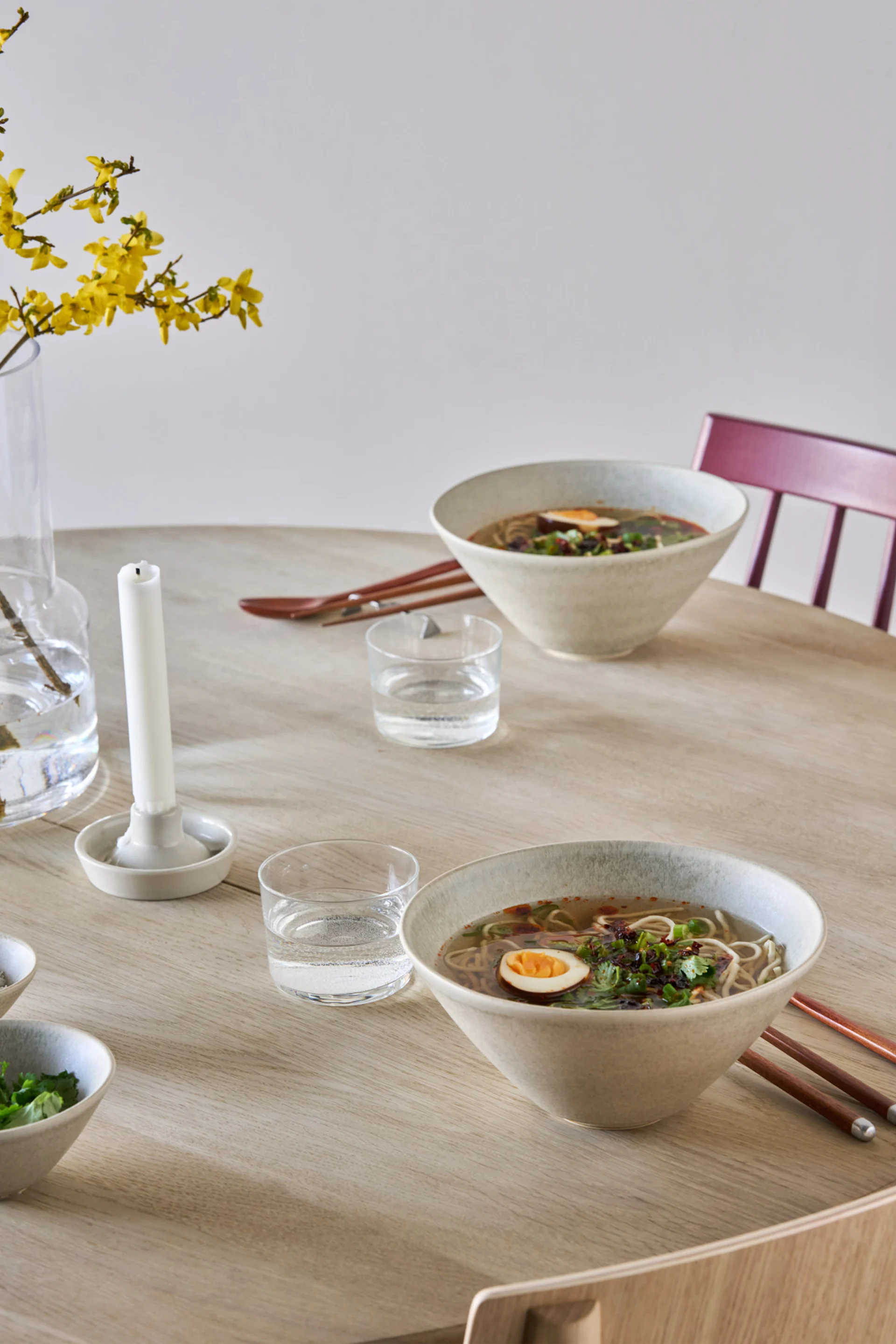 Dos tazones de ramen con huevos y guarnición sobre una mesa de madera clara, con vasos de agua y flores amarillas en un jarrón.