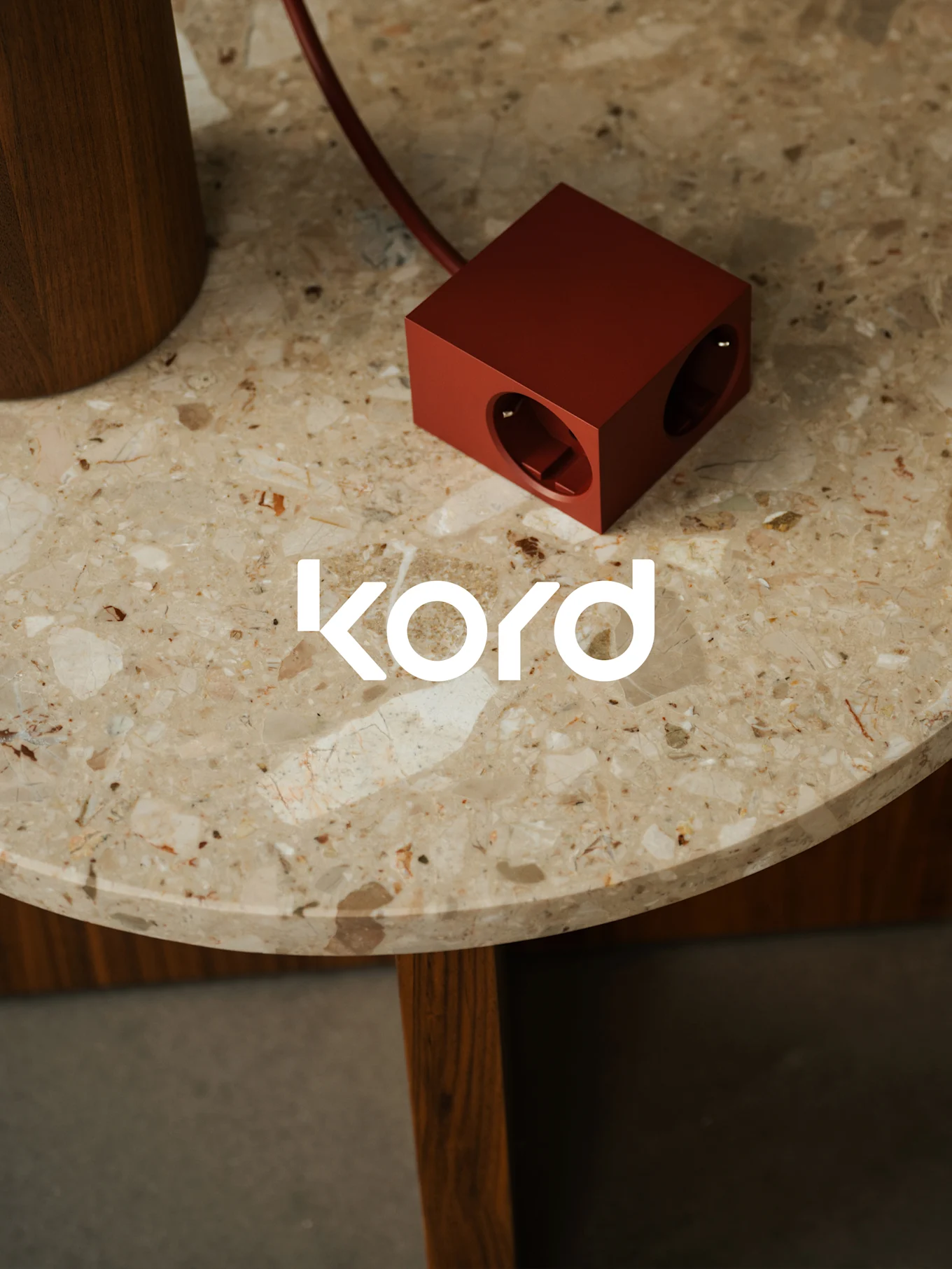 Una regleta de corriente de color rojo oscuro y forma cúbica con dos tomas visibles descansa sobre una mesa redonda de mármol, con el logo blanco "kord".
